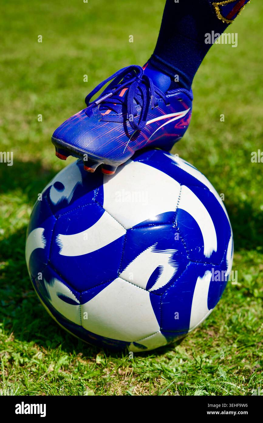 Nahaufnahme eines Fußes eines Fußballspielers mit blauem Schuh, der auf einem blau-weißen Fußball auf grünem Gras im Freien liegt Stockfoto