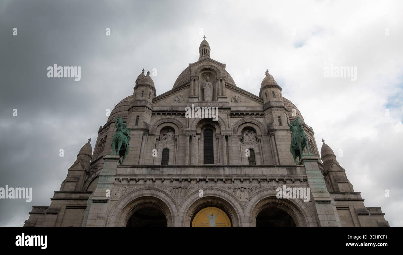 Flacher Blick auf die weiße Travertinfassade der Basilika Sacré-Cœur unter einem dramatischen bewölkten Himmel, Montmartre, Paris, Frankreich, Frühjahr 2026. Stockfoto
