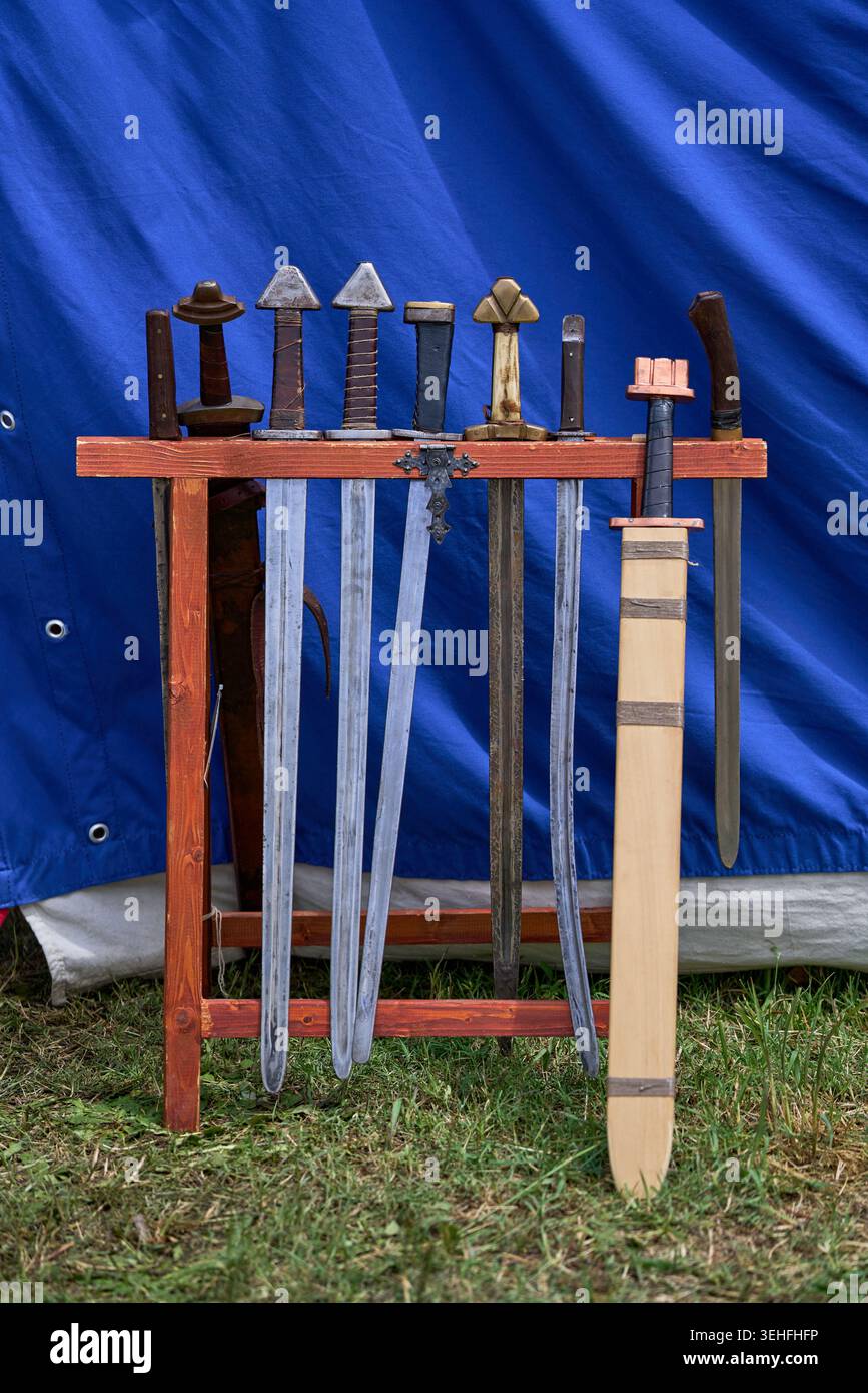 Historische Nachstellungen von Waffen, antike Klingen und eine hölzerne Debe stehen draußen auf einem Regal vor einem blauen Zelt Stockfoto