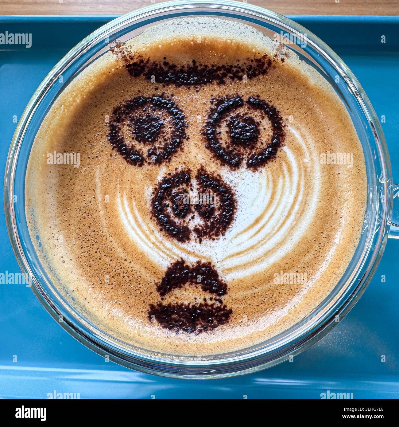 Lustiges Gesicht mit Kakaopulver auf einem Cappuccino-Kaffee, serviert in einer Glasschale auf einem blauen Tablett, von oben gesehen. - Smartphone-aufgenommenes Stockfoto