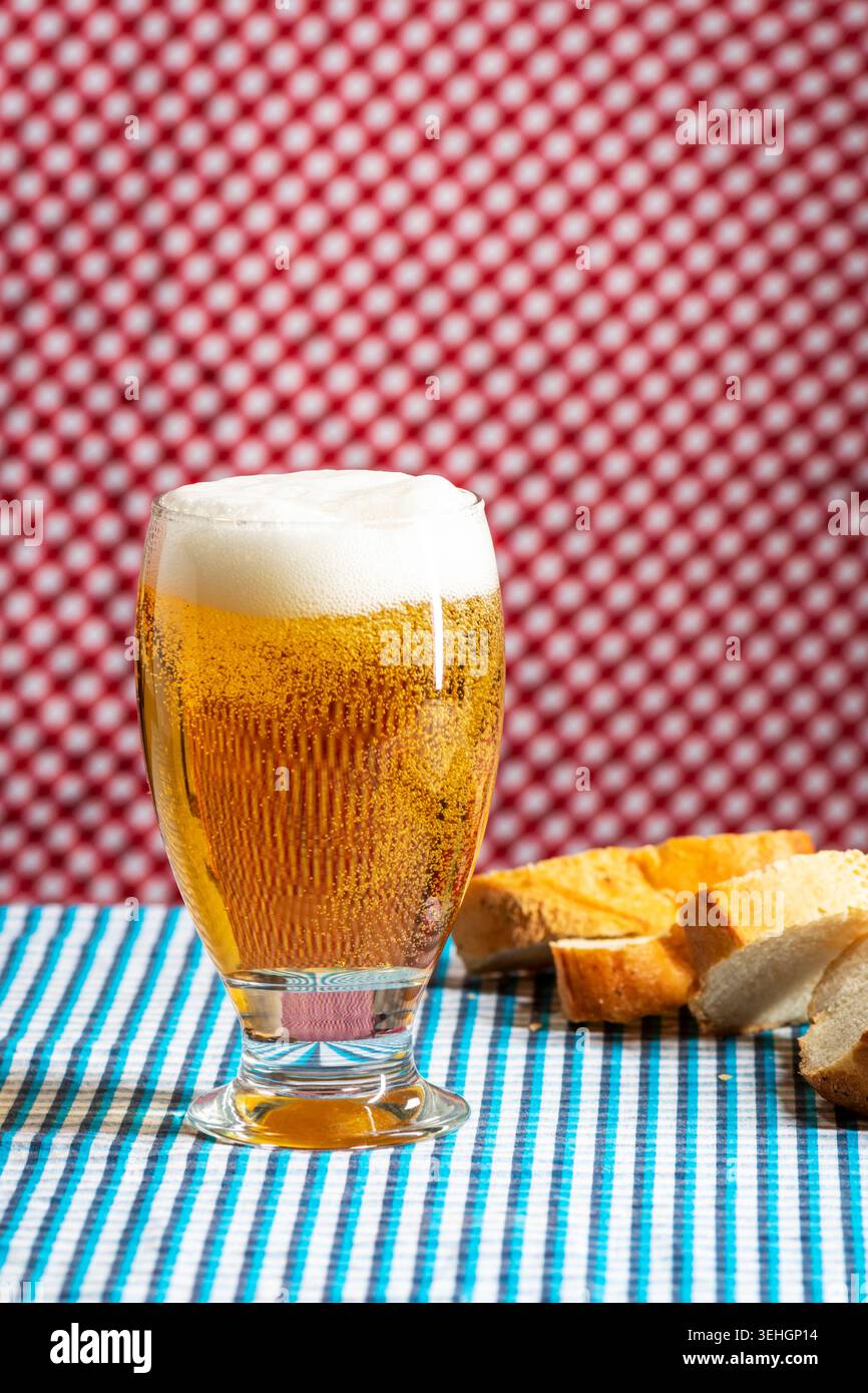 Goldenes Bier mit cremigem Schaum, serviert neben rustikalem Brot auf einer farbenfrohen Tischdecke im bayerischen Stil. Traditionelle Oktoberfestatmosphäre Stockfoto