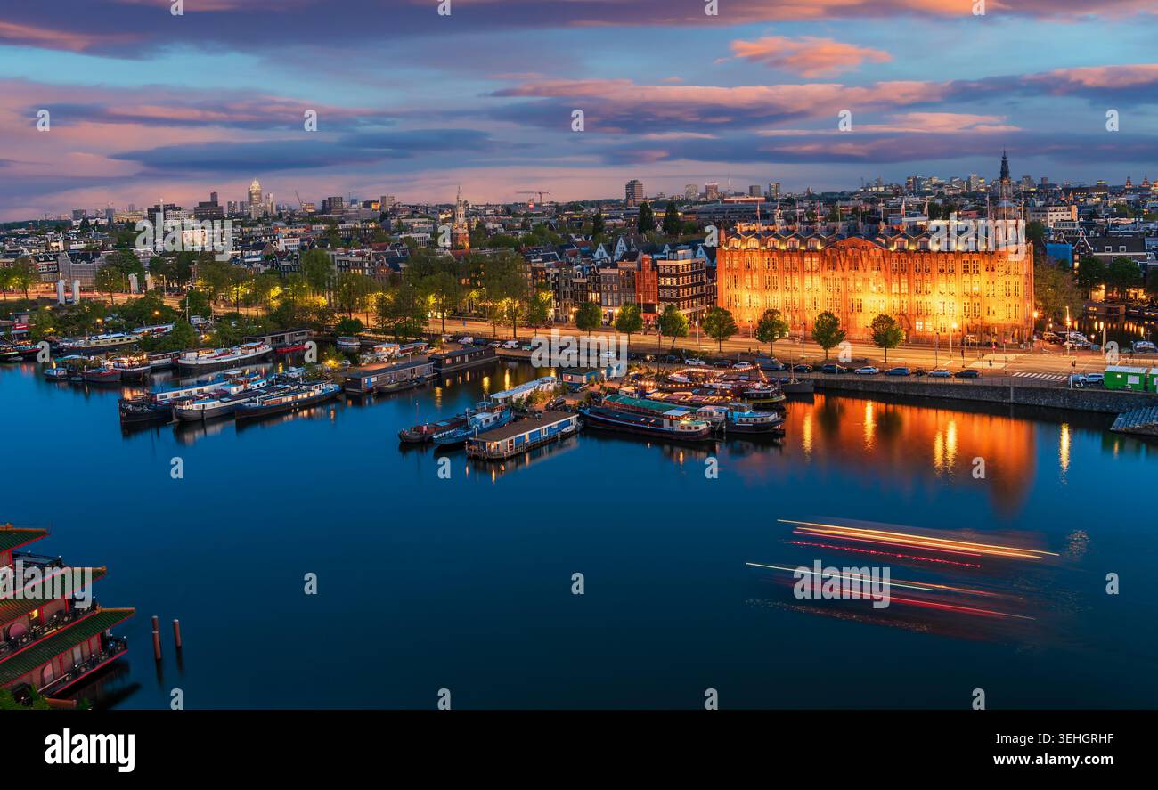 Blaues Stadtbild der Amsterdamer Uferpromenade mit leuchtender historischer Architektur, ruhigen Reflexen und dramatischem Abendhimmel. Stockfoto