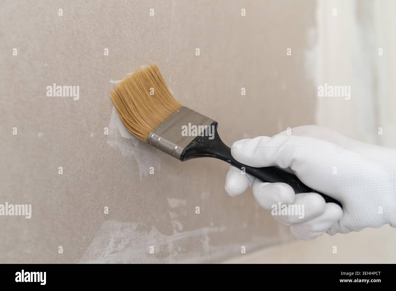 Eine Hand, die einen weißen Handschuh trägt, trägt mit einem hochwertigen Pinsel die Lackierung auf eine glatte Trockenbauoberfläche auf. Stockfoto