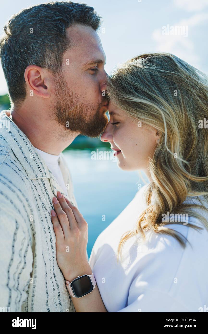 Ein Paar steht nahe am Wasser und genießt einen sonnigen Tag. Der Mann küsst die Stirn der Frau, während sie sich in ihn lehnt und ihre Verbindung mit mir zeigt Stockfoto
