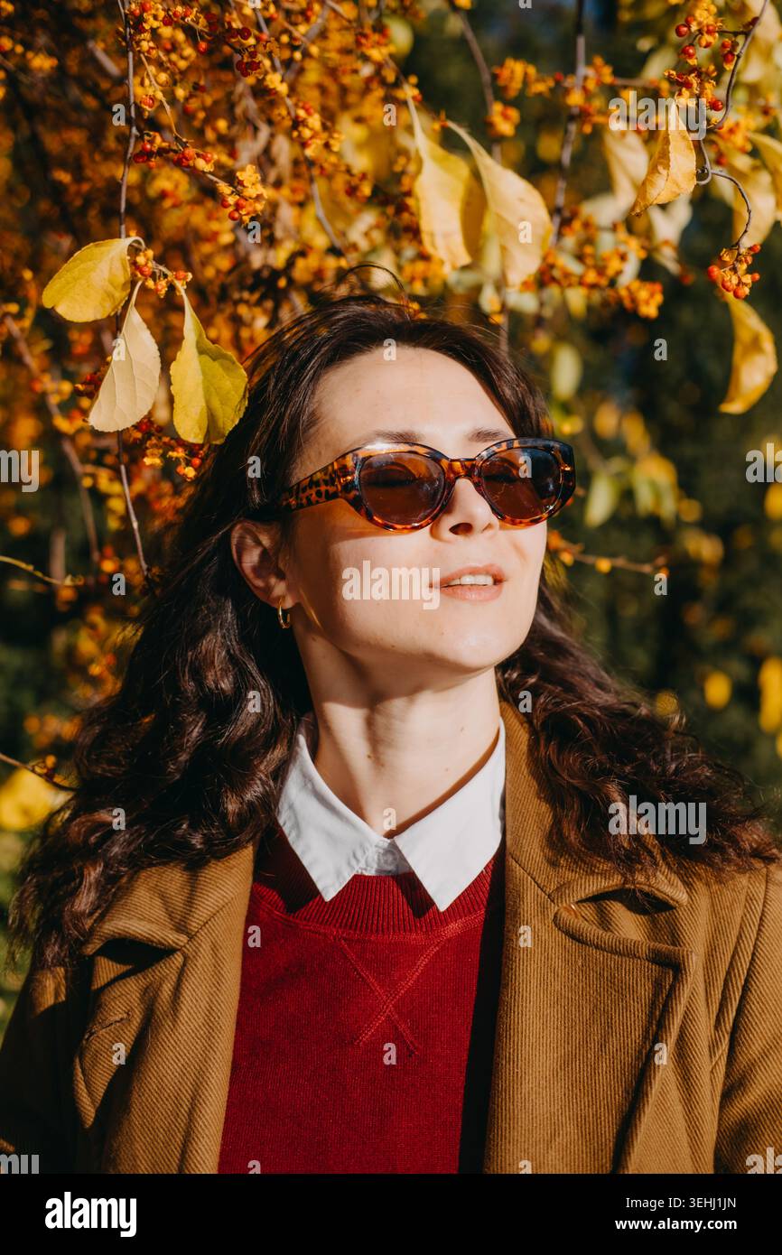 Frau posiert draußen mit Herbstzweigen, während sie eine Sonnenbrille trägt. Sonnenschutz im Herbst, Kampagne für uv-Sicherheit und Lifestyle, Übergangszeit Stockfoto