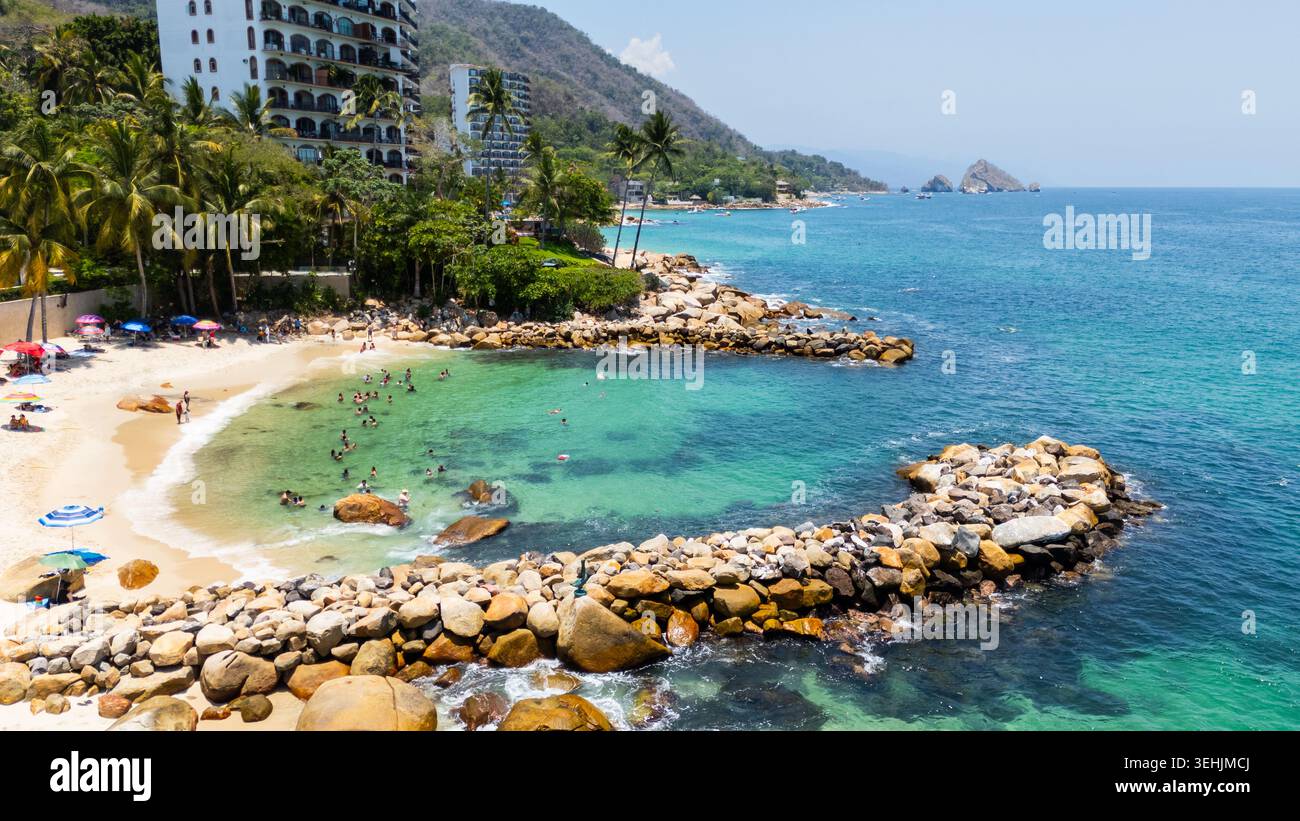 Aus der Vogelperspektive auf Playa Esmeralda mit Menschen, die im türkisfarbenen Wasser schwimmen, felsigen Wellenbrechern und Strandresorts in Puerto Vallarta, Jalisco, Mexiko. Stockfoto