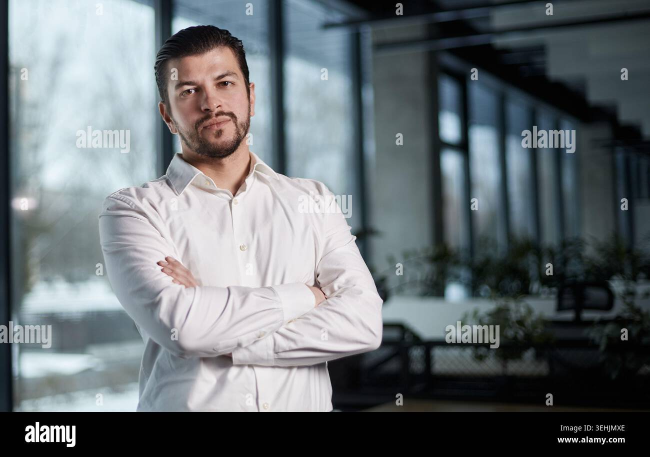 Ein Mann in einem weißen Hemd steht mit überkreuzten Armen vor einem Fenster Stockfoto