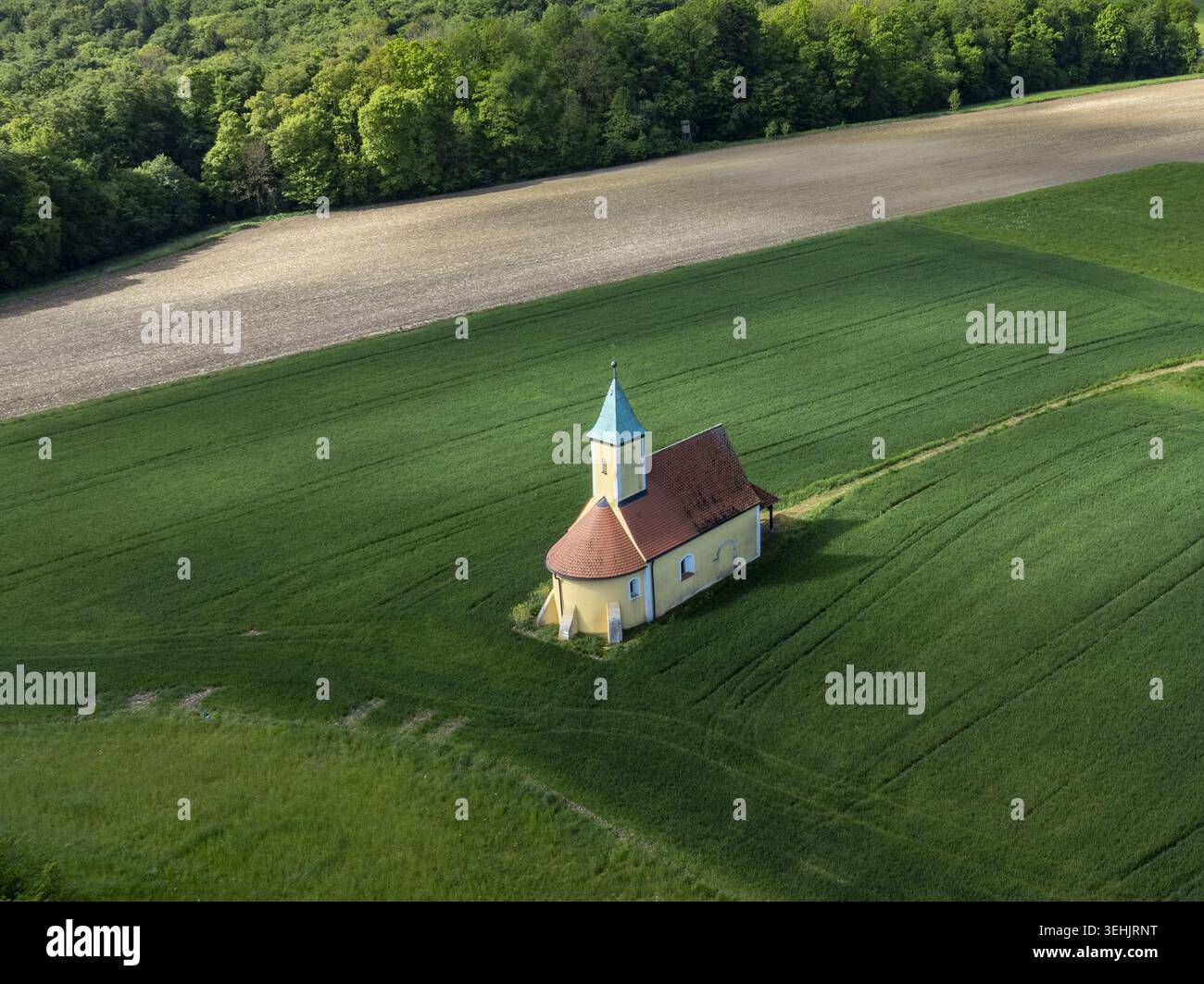 Aus der Vogelperspektive auf eine kleine historische Kapelle mit rotem Dach und grünem Turm, die inmitten üppiger grüner Felder in der Nähe eines walddeutschen Waldes steht Stockfoto