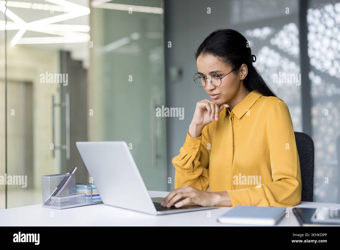 Junge Geschäftsfrau mit Brille und gelbem Hemd, die sich auf ihre Arbeit konzentriert, auf einem Laptop an einem modernen Schreibtisch schreibt und Hingabe und Effizienz verkörpert Stockfoto