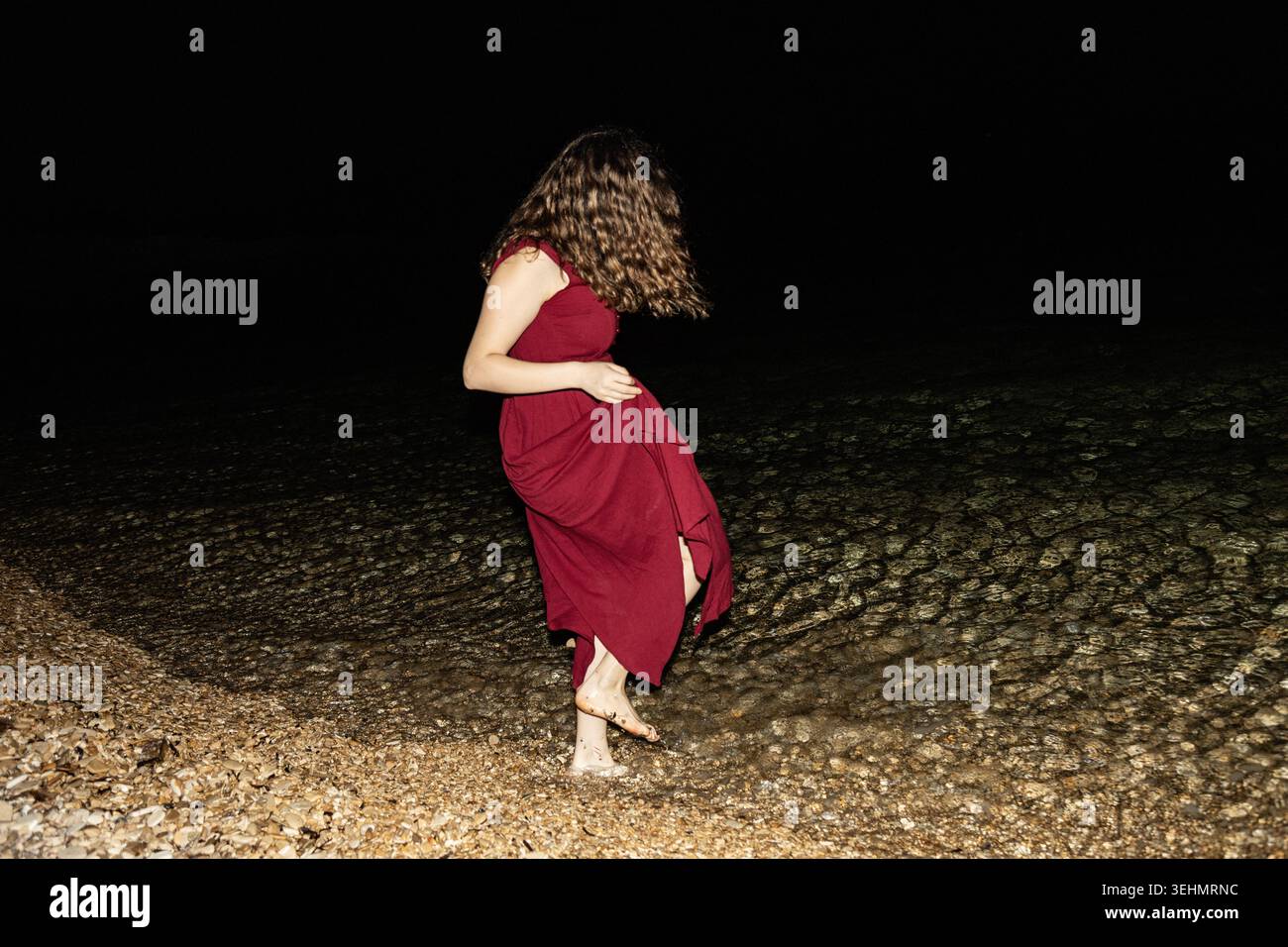 Eine Frau in einem fließenden roten Kleid tritt barfuß in flachem Wasser an einem Kiesstrand, beleuchtet durch Blitz vor einem dunklen Horizont und schafft ein stimmungsvolles Kino Stockfoto