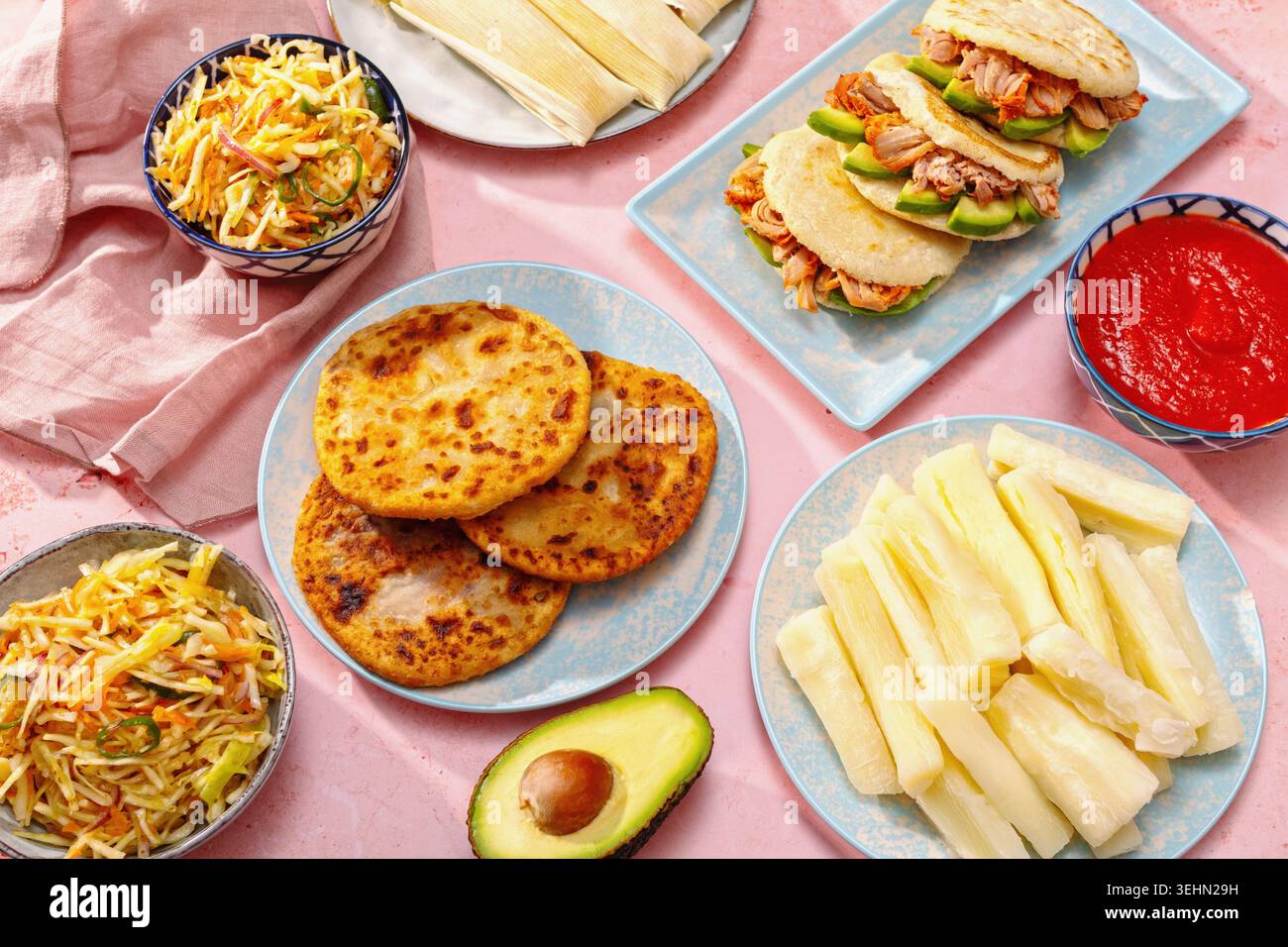 Tischbild mit lateinamerikanischem Familienessen mit Pupusas, Arepas, Curtido, Tajadas, Humitas, Yuca und Salsa auf einem farbenfrohen rosafarbenen Tisch, Tradition Stockfoto