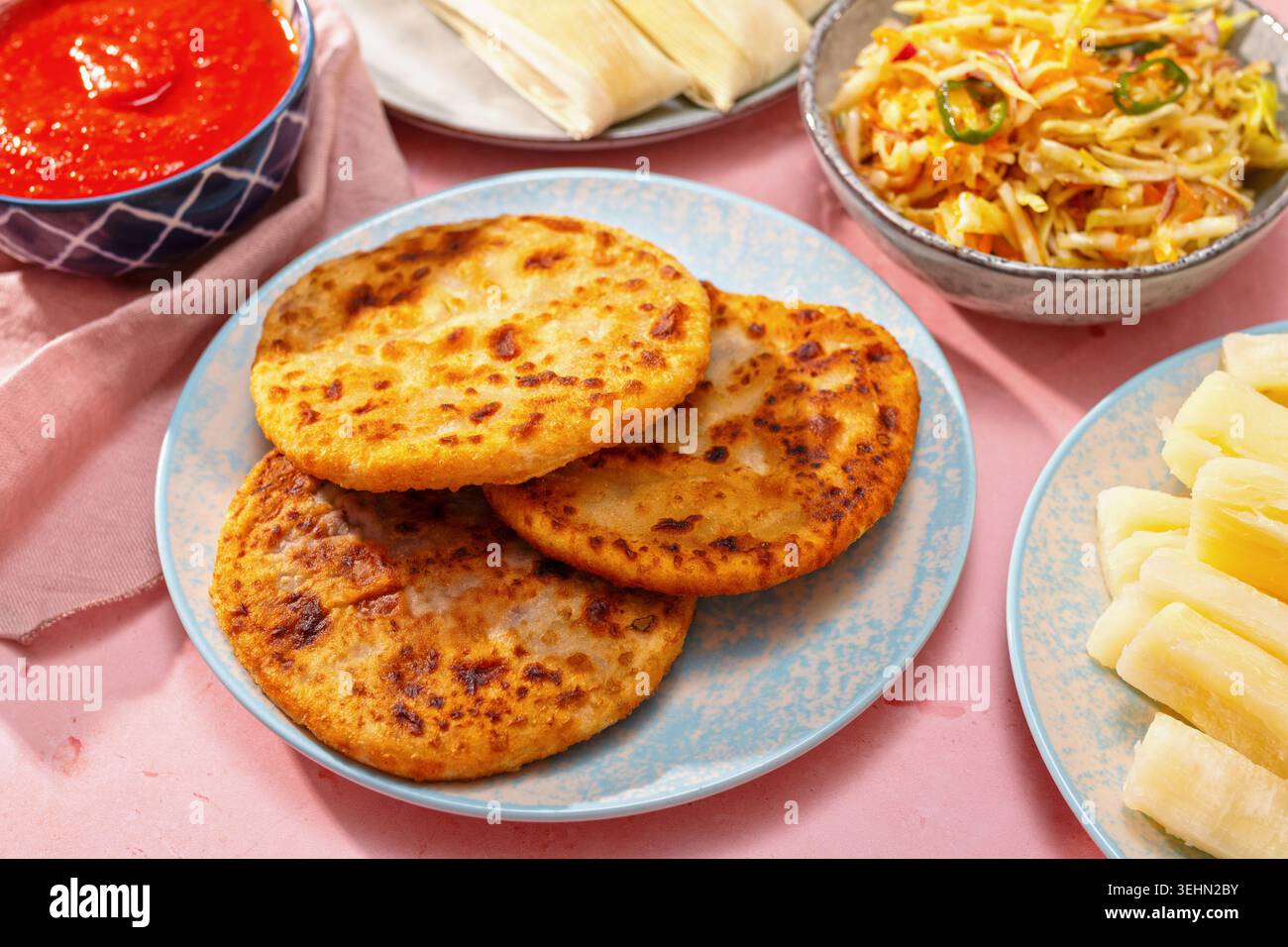 Pupusas auf einem blauen Teller serviert mit Curtido, Salsa roja, Yuca und Humitas auf rosa Hintergrund, die ein traditionelles salvadorianisches Essen und Central A zeigen Stockfoto