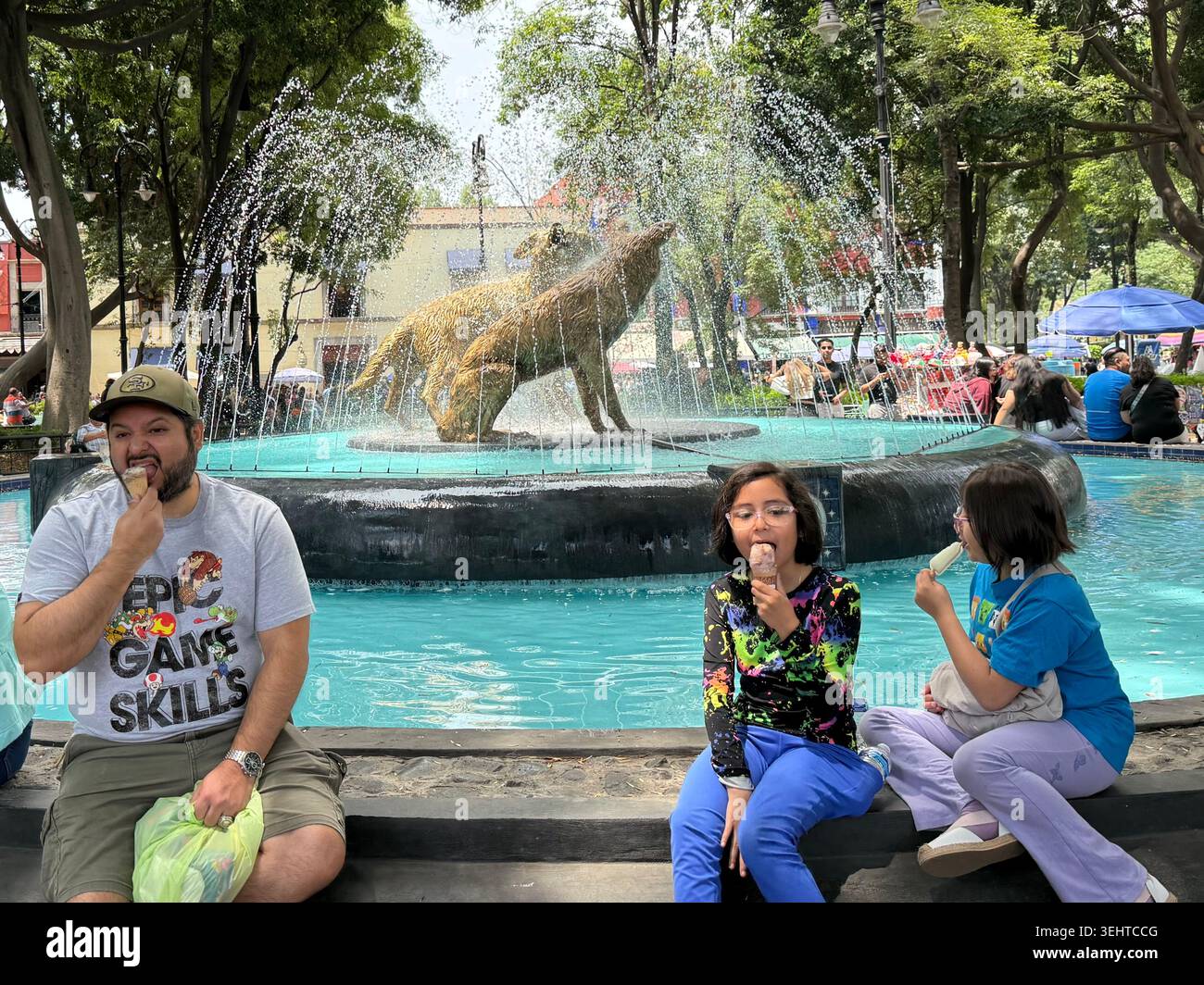 Die Menschen essen Eis neben der Fuente de los Coyotes in Jardín Centenario, einem beliebten öffentlichen Platz in Coyoacán, Mexiko-Stadt - Smartphone-aufgenommenes Stockfoto