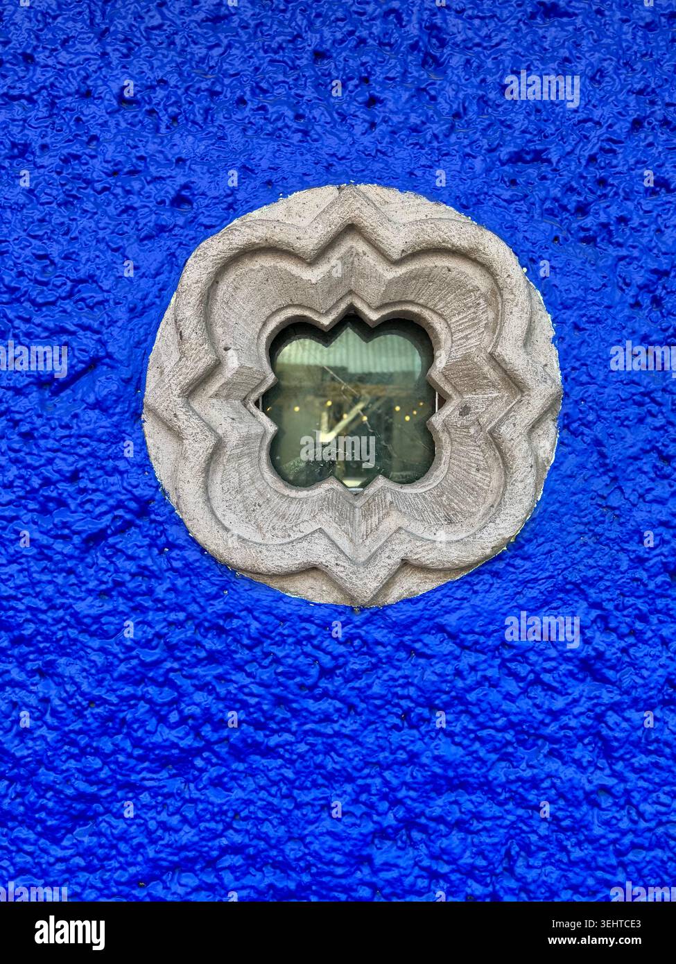 Dekoratives Fenster mit Steinrahmen in einer leuchtend blau strukturierten Wand in Coyoacán, Mexiko-Stadt, Mexiko - Smartphone-aufgenommenes Stockfoto