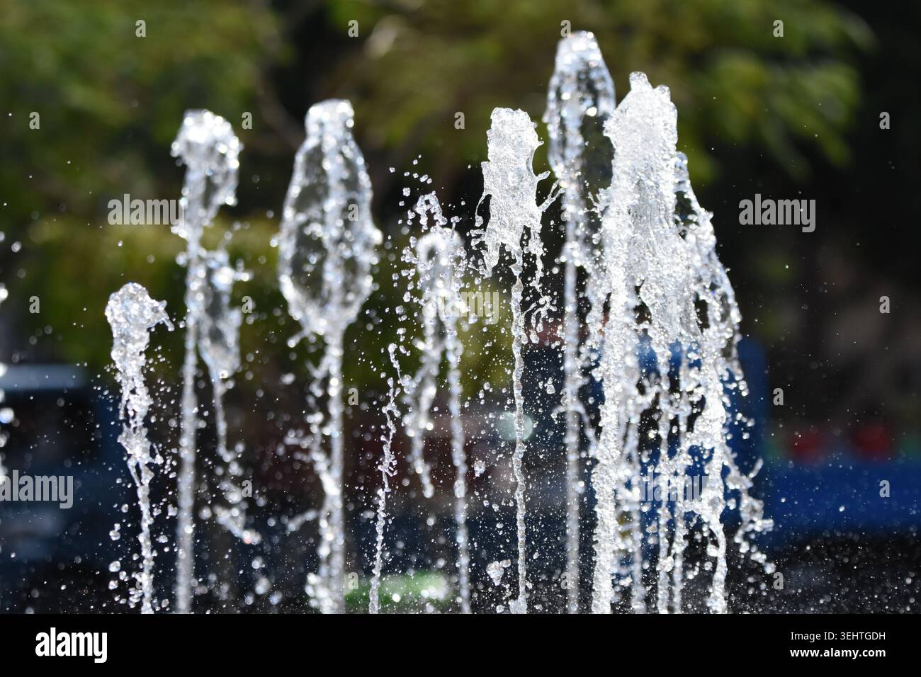 Wasser spritzt aus mehreren Wasserstrahlen eines Springbrunnens und bildet eine fesselnde Tropfenanzeige vor einem hellen sonnigen Himmel in einem lebhaften Stadtpark. Personen en Stockfoto