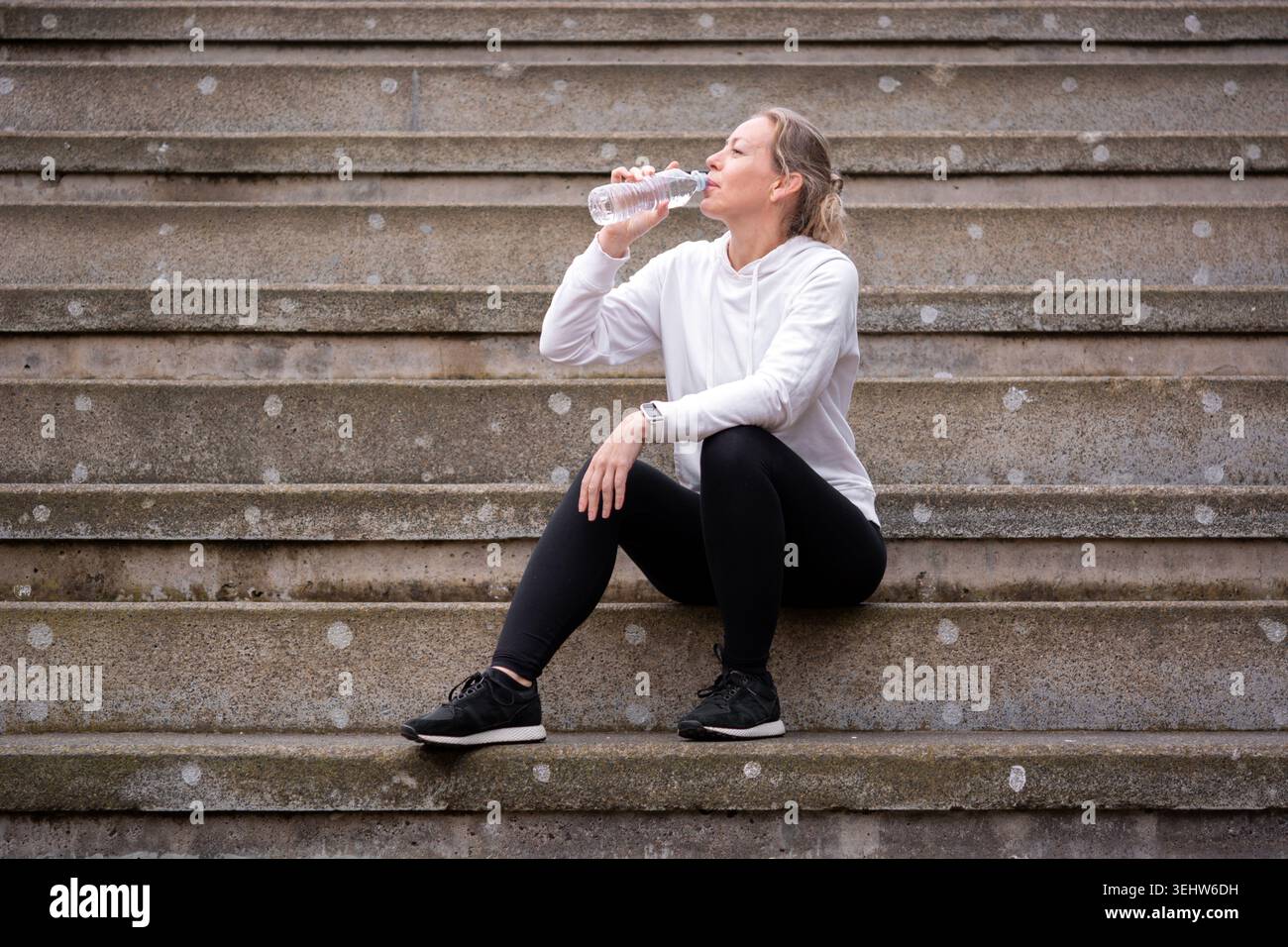 Aktive Frau, die auf Betontreppen ruht und nach dem Training im Freien Wasser trinkt. Gesundes Lifestyle- und Fitnesskonzept mit sportlicher Kleidung und nat Stockfoto