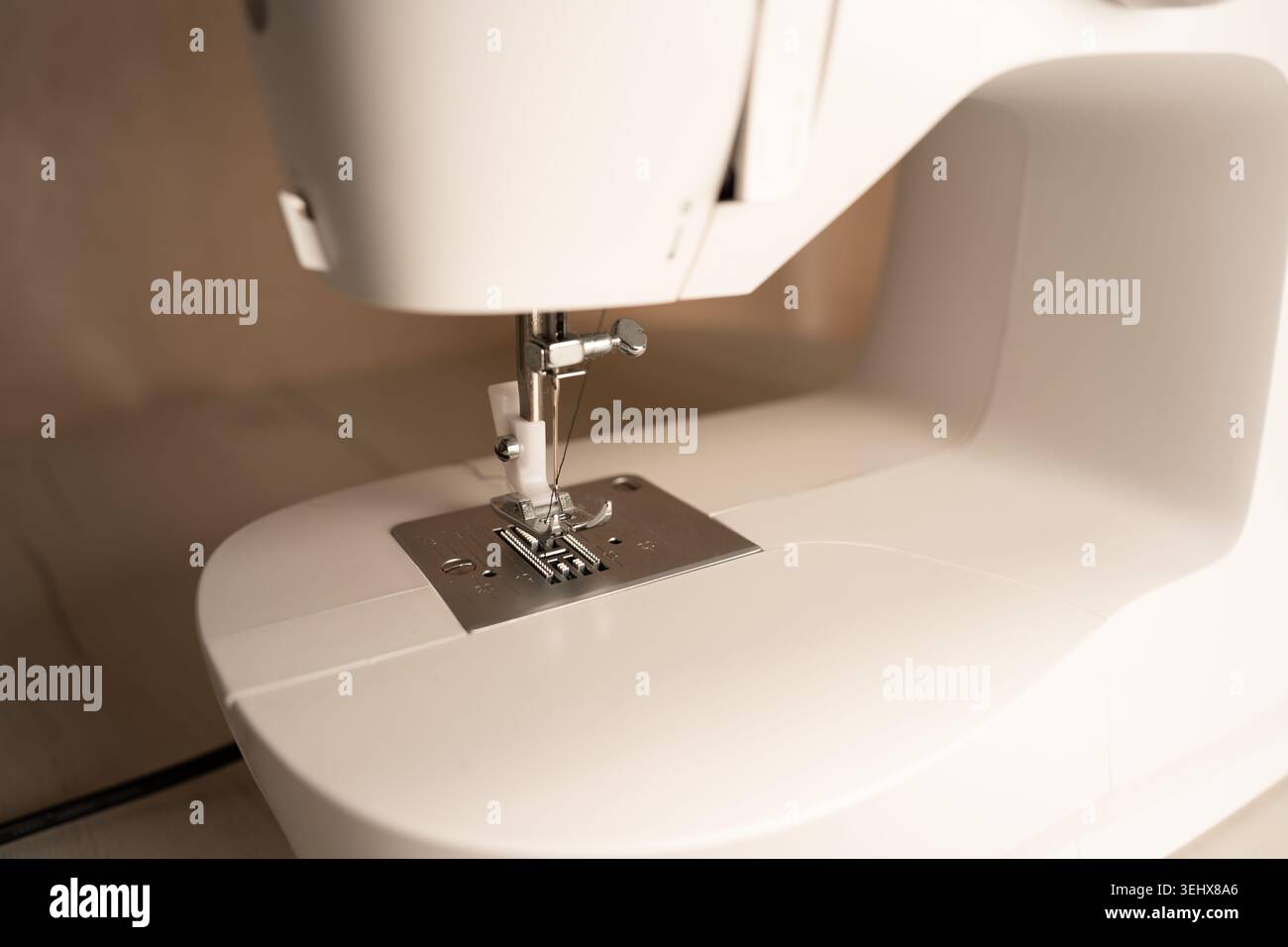Nahaufnahme einer modernen weißen Nähmaschine mit Nähfuß, Nadel und Chromfüßhunden auf einer cremefarbenen Tischplatte mit warmem Weichfokus, min Stockfoto