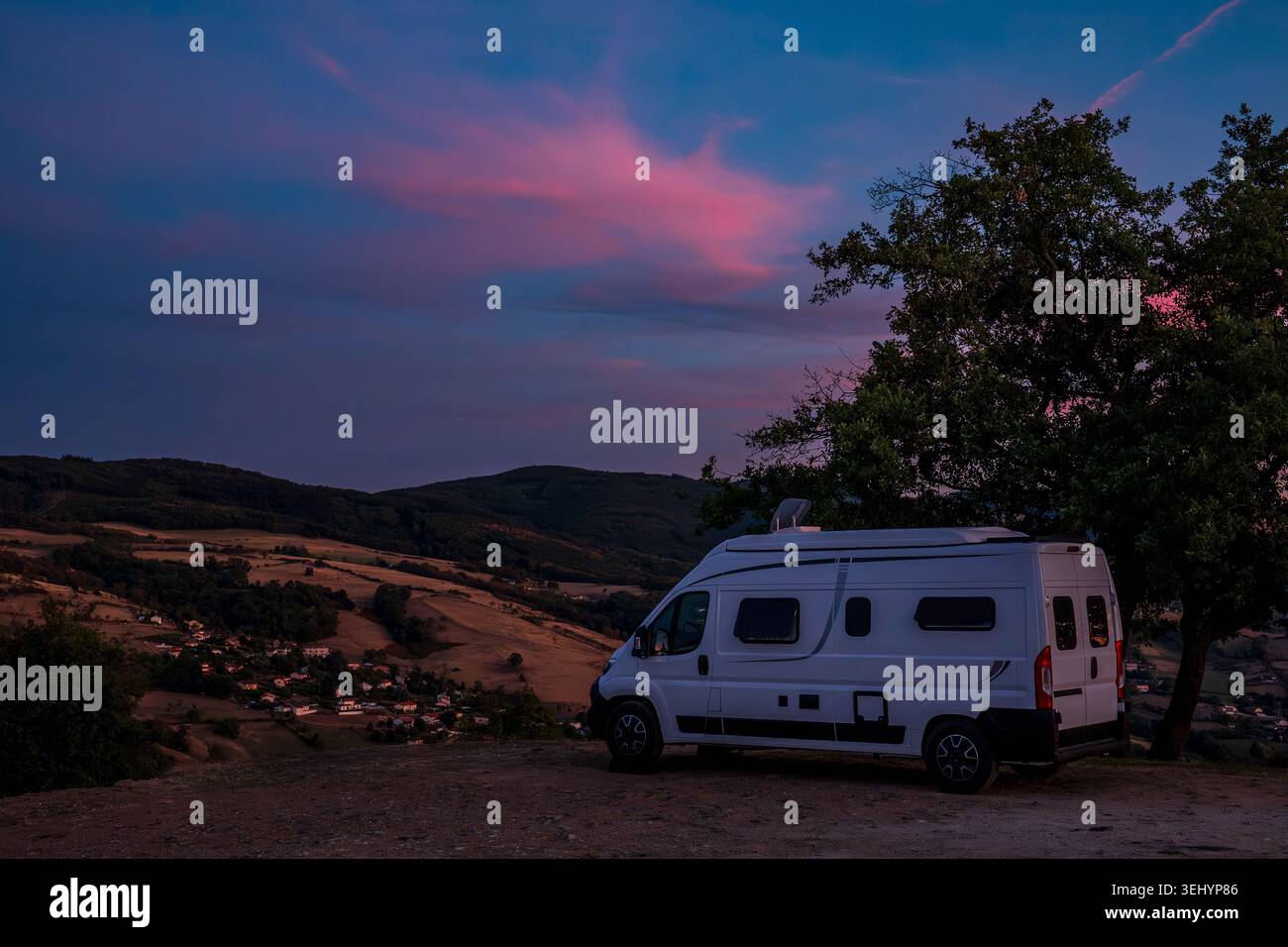 Ein weißer Wohnmobil steht auf einem malerischen Hügel neben einem großen Baum während der Dämmerung. Im Hintergrund verblassen die Hügel und Dörfer Beaujolais in dar Stockfoto