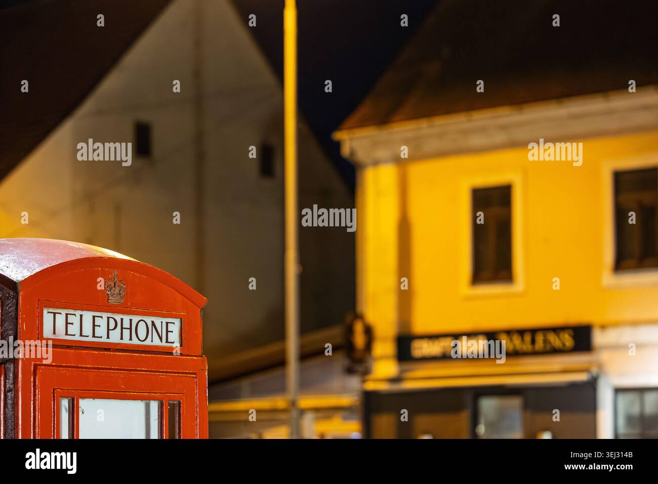 Vinkovci, Kroatien - 22. März 2026: Eine klassische rote Telefonkabine steht nachts auf einer ruhigen Stadtstraße, die in warmes gelbes Licht aus dem nahegelegenen Buil getaucht ist Stockfoto