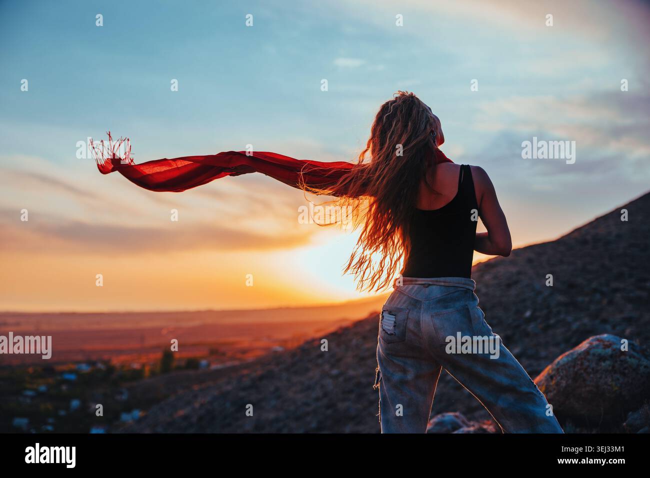 Junge blonde Frau mit langen Haaren und einem fließenden roten Schal, die bei Sonnenuntergang in den Bergen steht Stockfoto