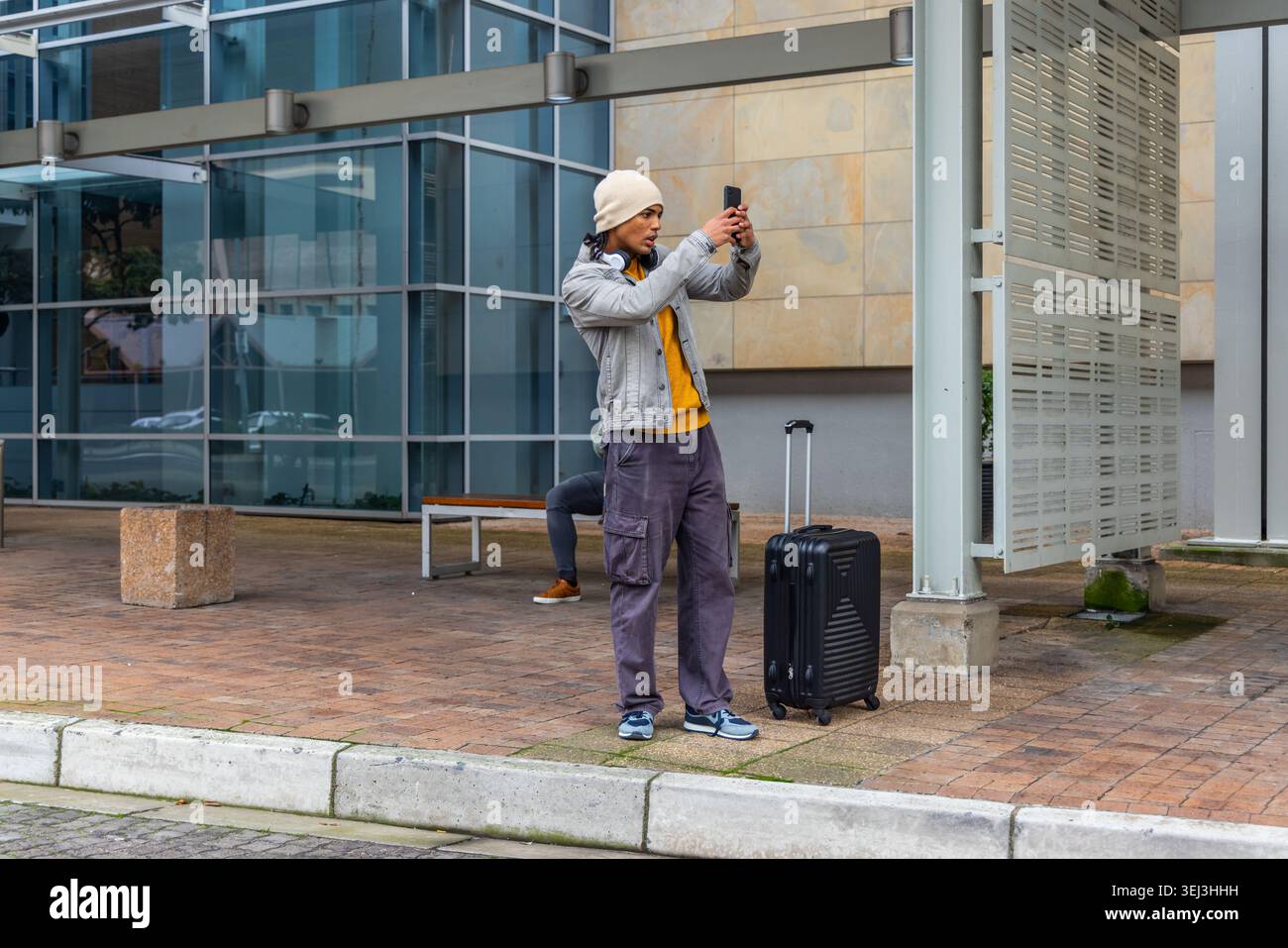 Ein Mann, der das Smartphone auf der plaza überprüft, mit Beanie und Jeansjacke, mit Rollkoffer Stockfoto