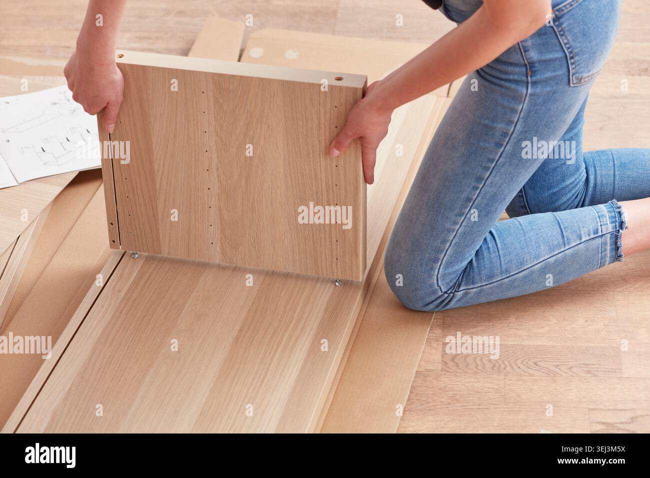 Eine Person stellt sorgfältig ein Holzmöbel auf dem Boden zusammen, umgeben von Verpackungsmaterialien, in einem gut beleuchteten Raum, der mit natürlichem L gefüllt ist Stockfoto