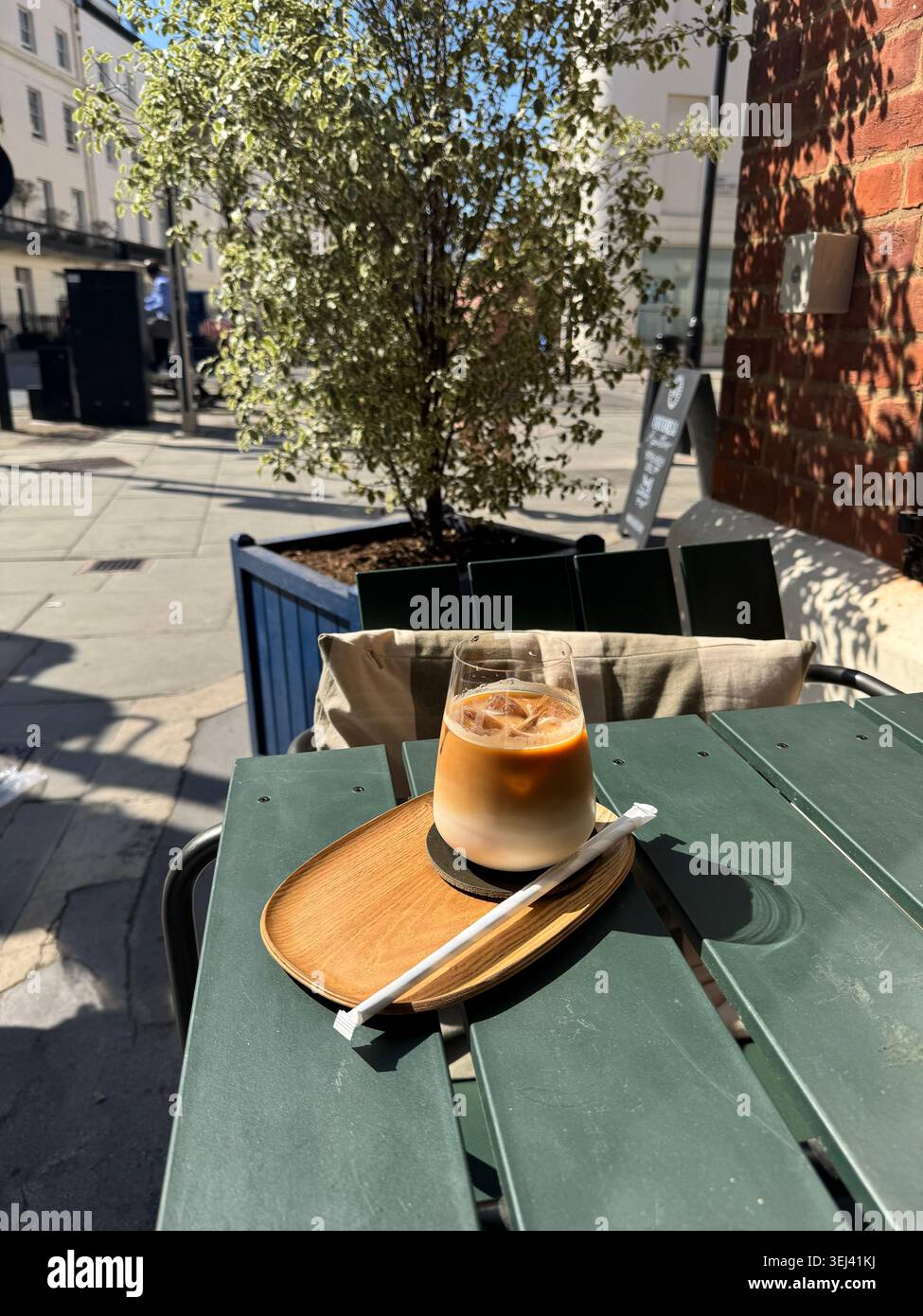 Ein Eiskaffee auf einem kleinen Tablett serviert auf einem grünen Kaffeetisch mit grünem Baum und sonniger Stadtstraße im Hintergrund. - Smartphone-aufgenommenes Stockfoto