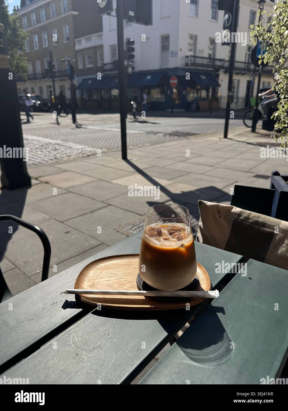 Ein Glas erfrischenden Eiskaffee Latte steht auf einem kleinen hölzernen Serviertablett auf einem dunkelgrünen Terrassentisch in einem Straßencafé in Belgravia, London. - Smartphone-aufgenommenes Stockfoto