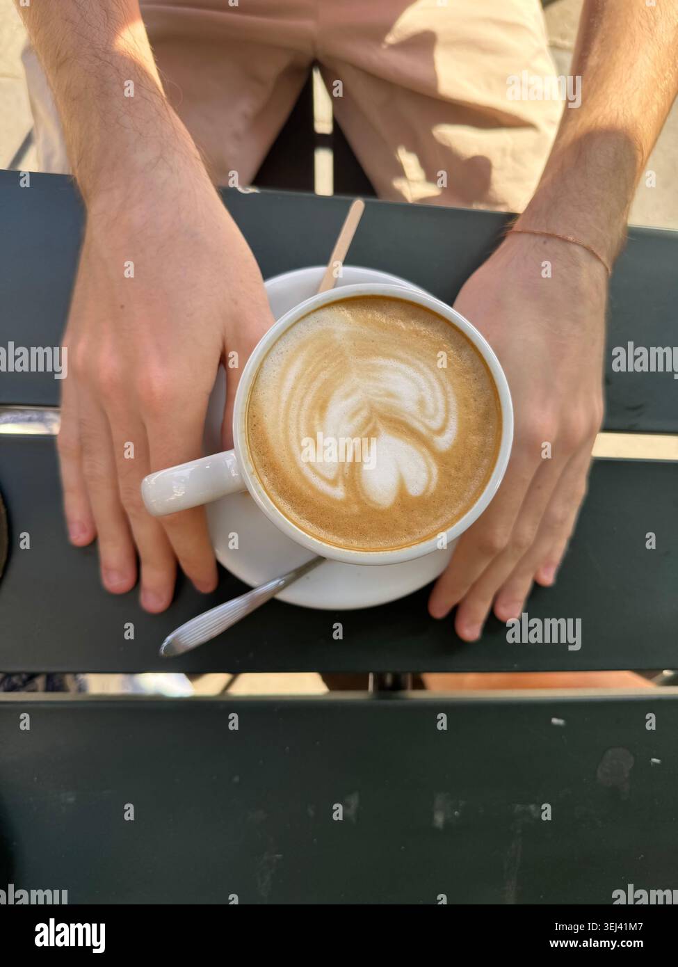 Blick von oben auf die Hände einer Person, die sanft um eine weiße Tasse Cappuccino mit Latte Art auf einem dunklen Terrassentisch in einem Londoner Café liegt. - Smartphone-aufgenommenes Stockfoto