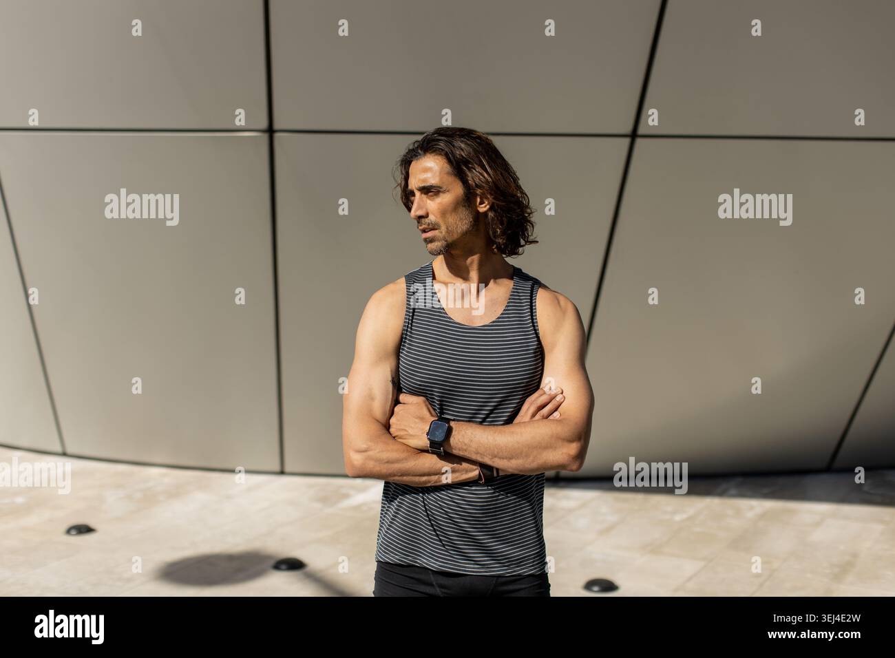 Ein Mann mit langen Haaren steht mit überkreuzten Armen und zeigt lässige Mode, fast schlankes, geometrisches Gebäudedesign unter hellem Sonnenlicht. Stockfoto