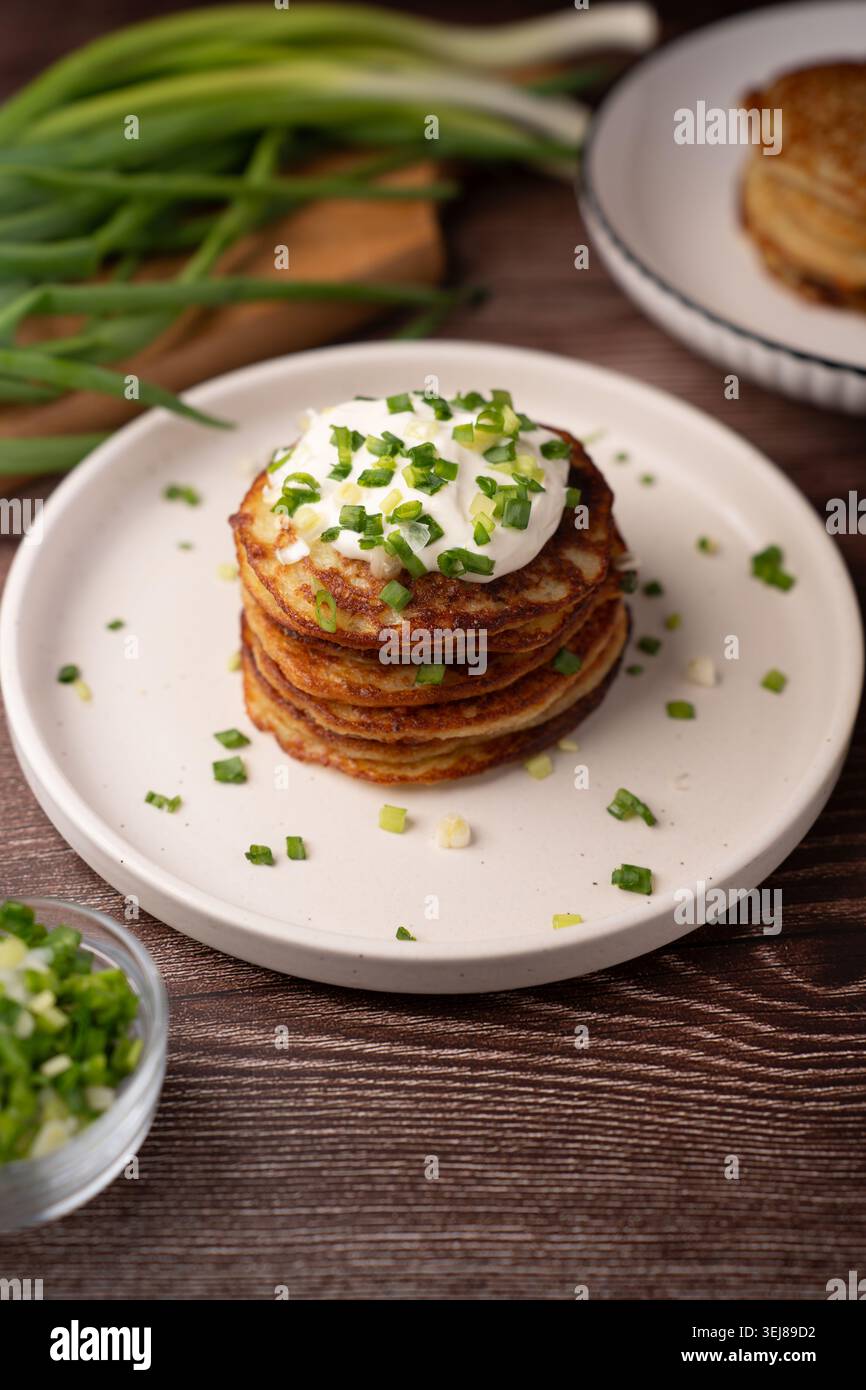 Stapel goldbrauner Kartoffelpfannkuchen mit cremiger Sauerrahm und hell gehacktem Schnittlauch auf einem weißen Teller, rustikaler Holztisch, frische Zwiebeln i Stockfoto