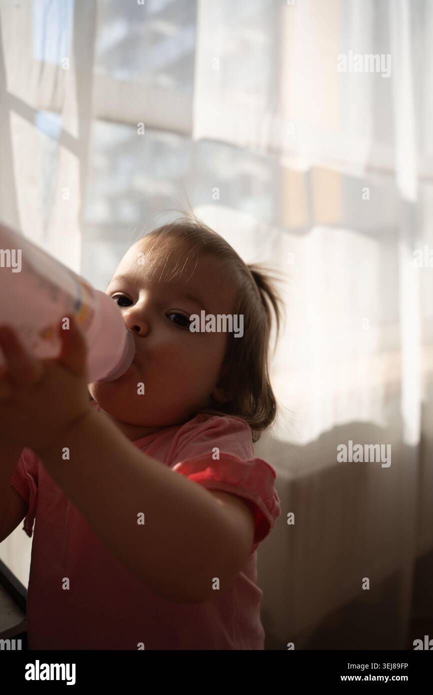 Nahaufnahme eines Kleinkindes, das aus einer rosa Flasche trinkt, in sanfter Morgenbeleuchtung durch transparente Vorhänge, eine warme, gemütliche Hausszene mit sanften Schatten Stockfoto