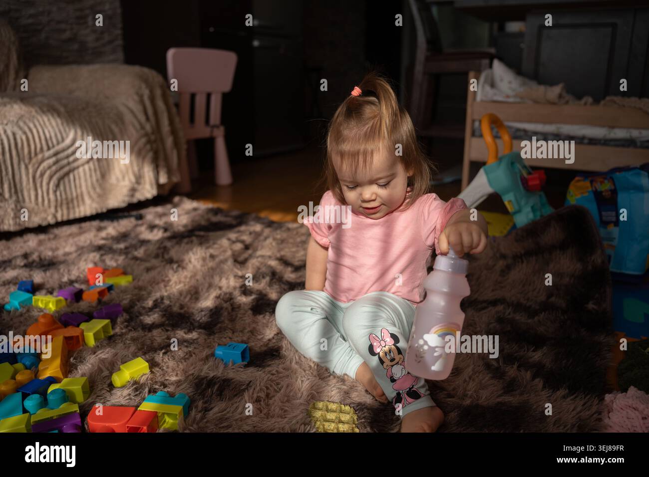 Das Kleinkind saß auf einem weichen braunen Teppich in einem sonnendurchfluteten Wohnzimmer, trug ein rosafarbenes T-Shirt und minnie-Leggings, hielt eine pastellfarbene Flasche zwischen bunten Blöcken und Stockfoto