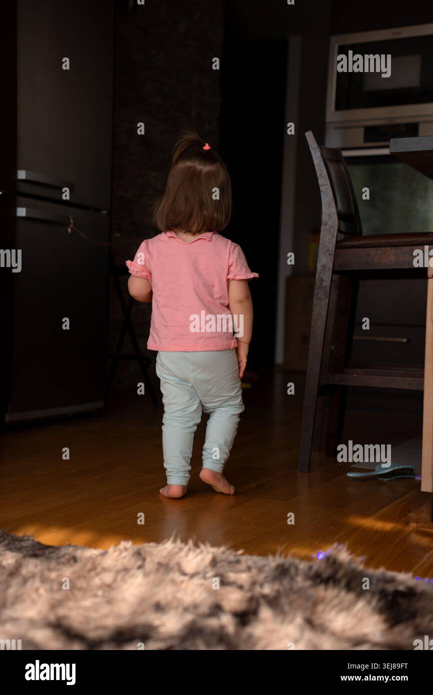 Rückansicht eines Kleinkindes in rosa Hemd und leichten Leggings, der barfuß über warmen Küchenboden aus Hartholz zu einem dunklen Eingang geht, weiches natürliches Licht Stockfoto