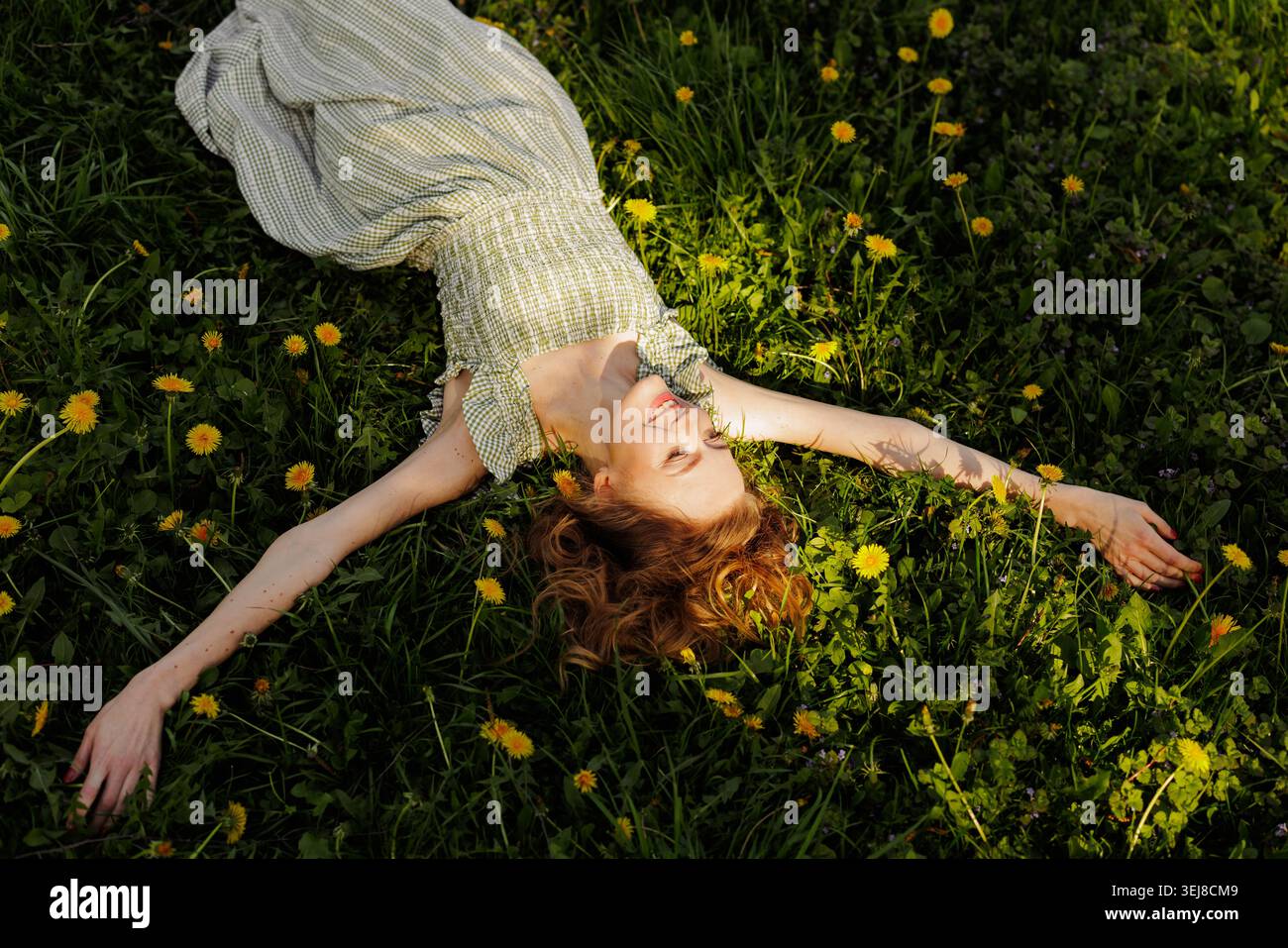 Eine junge Frau liegt auf einem grasbewachsenen Feld voller Löwenzahn und genießt einen sonnigen Tag, perfekt für Wellness, Lifestyle oder Naturprojekte. Stockfoto