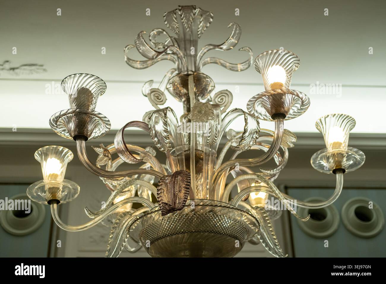 Der obere Teil einer durchdachten Stehlampe aus klarem Glas mit beleuchteten Lampen befindet sich unter einer weißen Decke in einem Innenbereich. Stockfoto