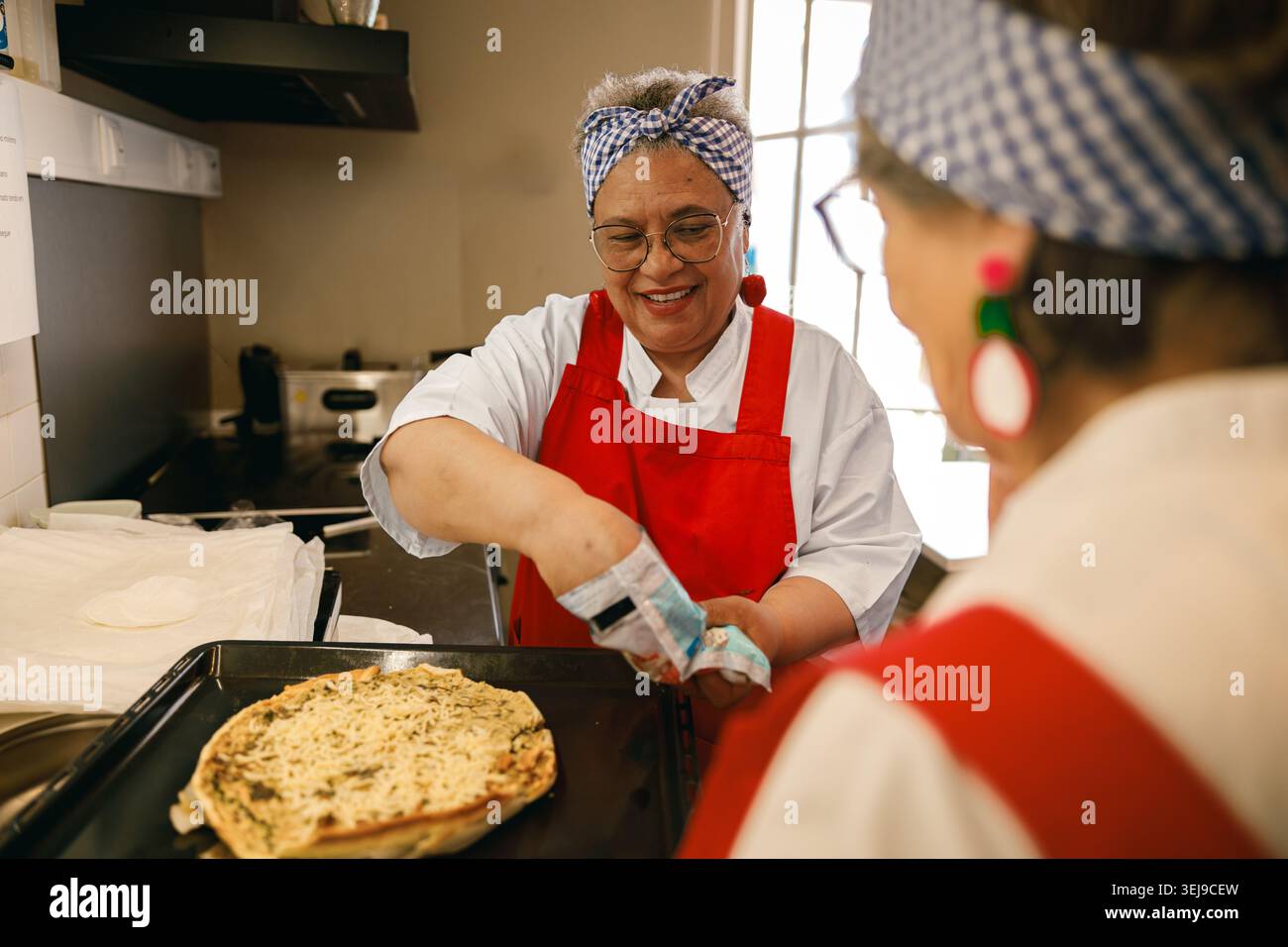 Lächelnde reife hispanische Frau, die Pizza in einer hellen Gemeinschaftsküche mit einer Freundin backt Stockfoto