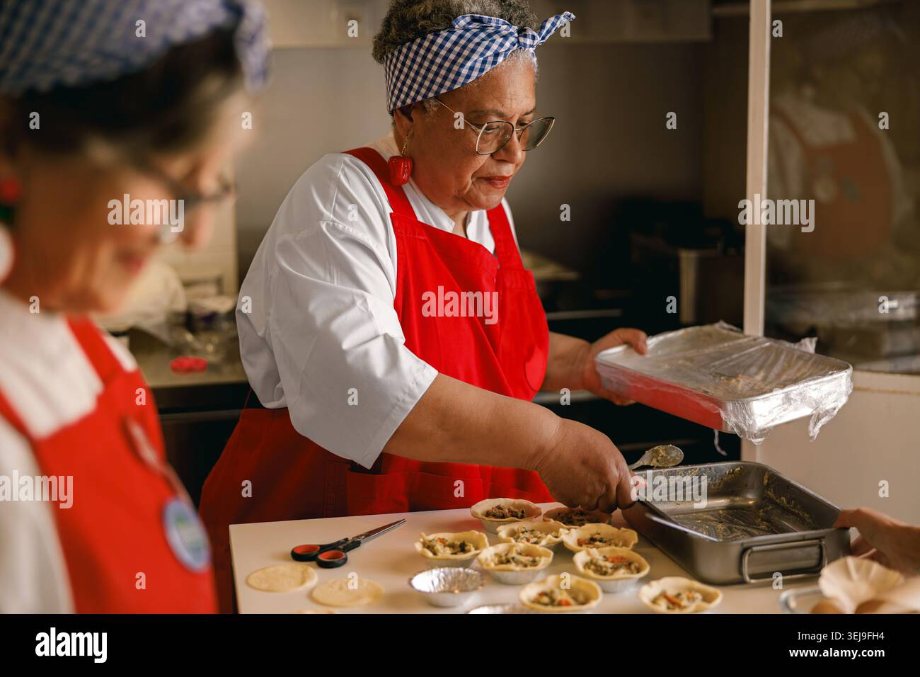 Ranghohe Afroamerikanerin, die in einer Gemeinschaftsküche Essen zubereitet, trägt rote Schürze und Stirnband. Stockfoto