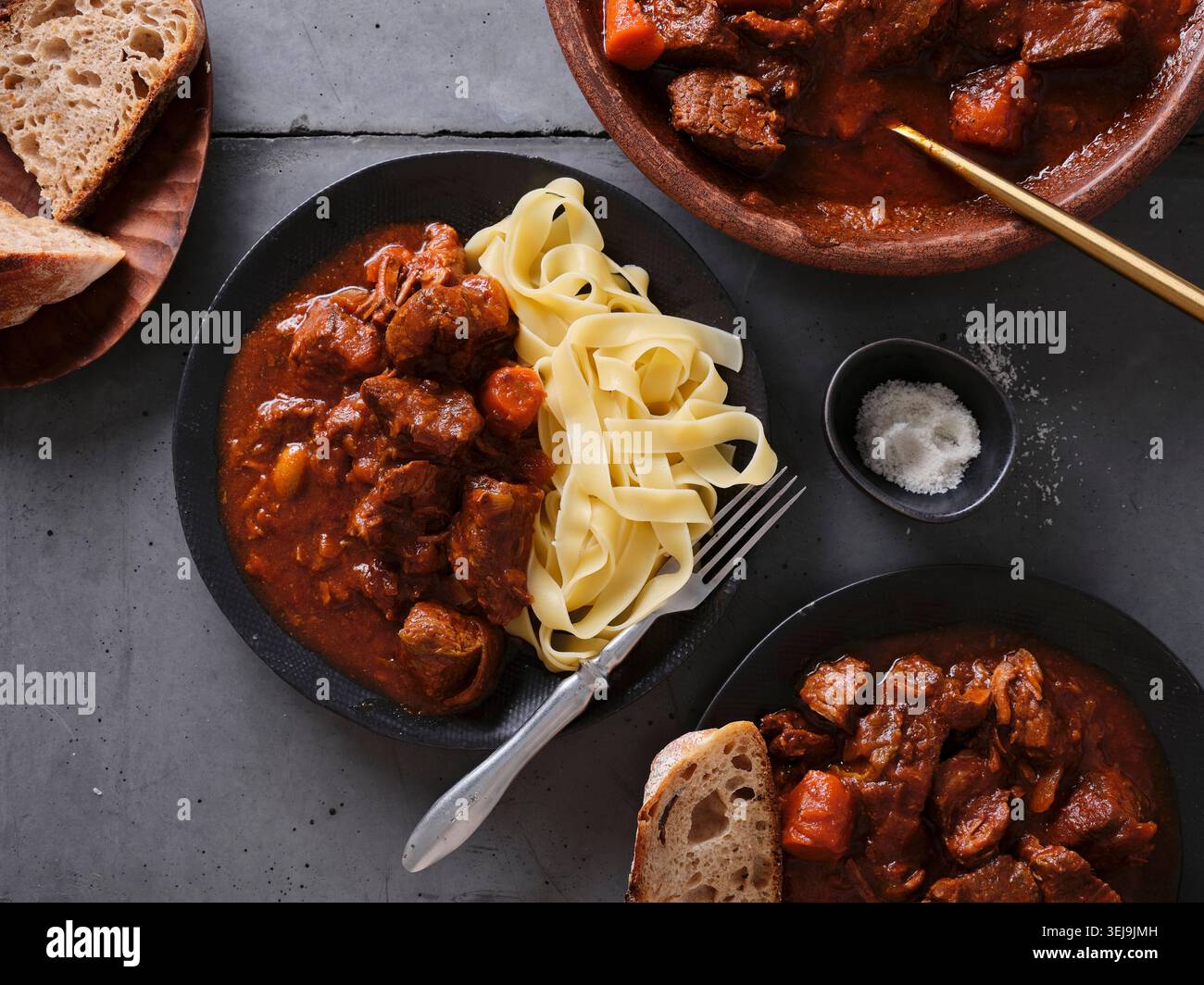 Langsam gekochtes, geriebenes Rindfleisch wird mit Nudeln überzogen und zusammen mit frischen Brotscheiben serviert. Die Umgebung zeigt einen Esstisch mit warmen Speisen und einem Casua Stockfoto