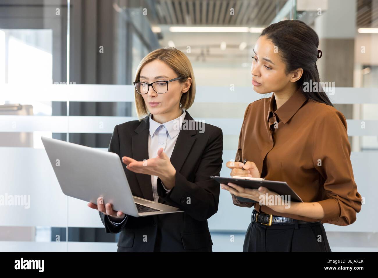 Zwei multirassische Geschäftsfrauen stehen in einem modernen Büro und diskutieren aktiv über ein neues Geschäftsprojekt, während eine einen Laptop hält und die andere Notizen macht Stockfoto