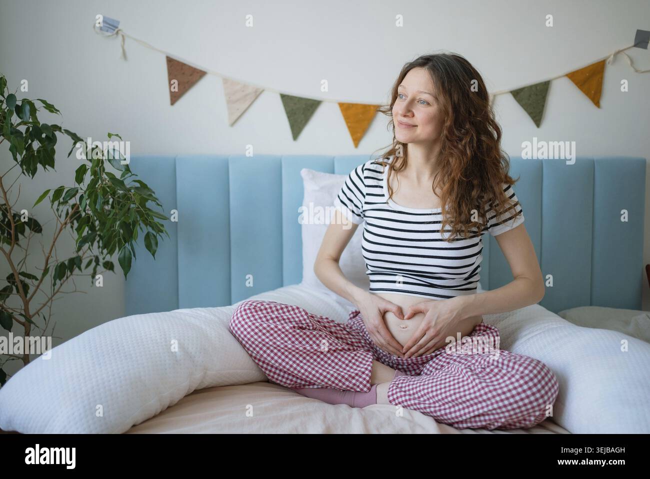 Eine schwangere Frau sitzt bequem im Bett, formt mit ihren Haenden ein Herz auf ihrem wachsenden Bauch, traeumt von der Mutterschaft zu Hause und geni Stockfoto