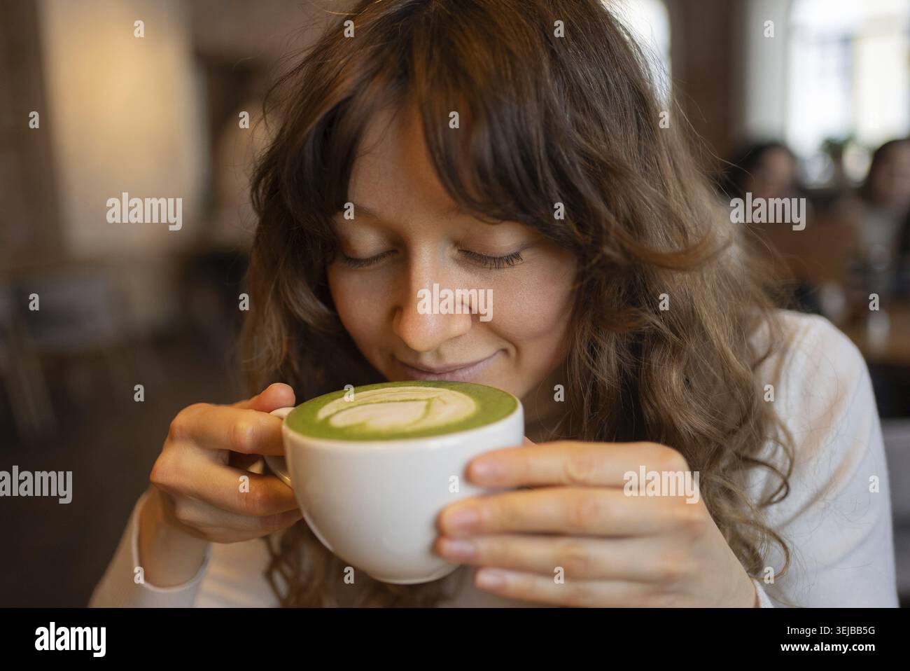 Eine junge Frau mit lockigen Haaren genießt den Duft eines Matcha Latte in einem warmen und einladenden Café und genießt einen Moment der Ruhe inmitten des belebten Atmos Stockfoto