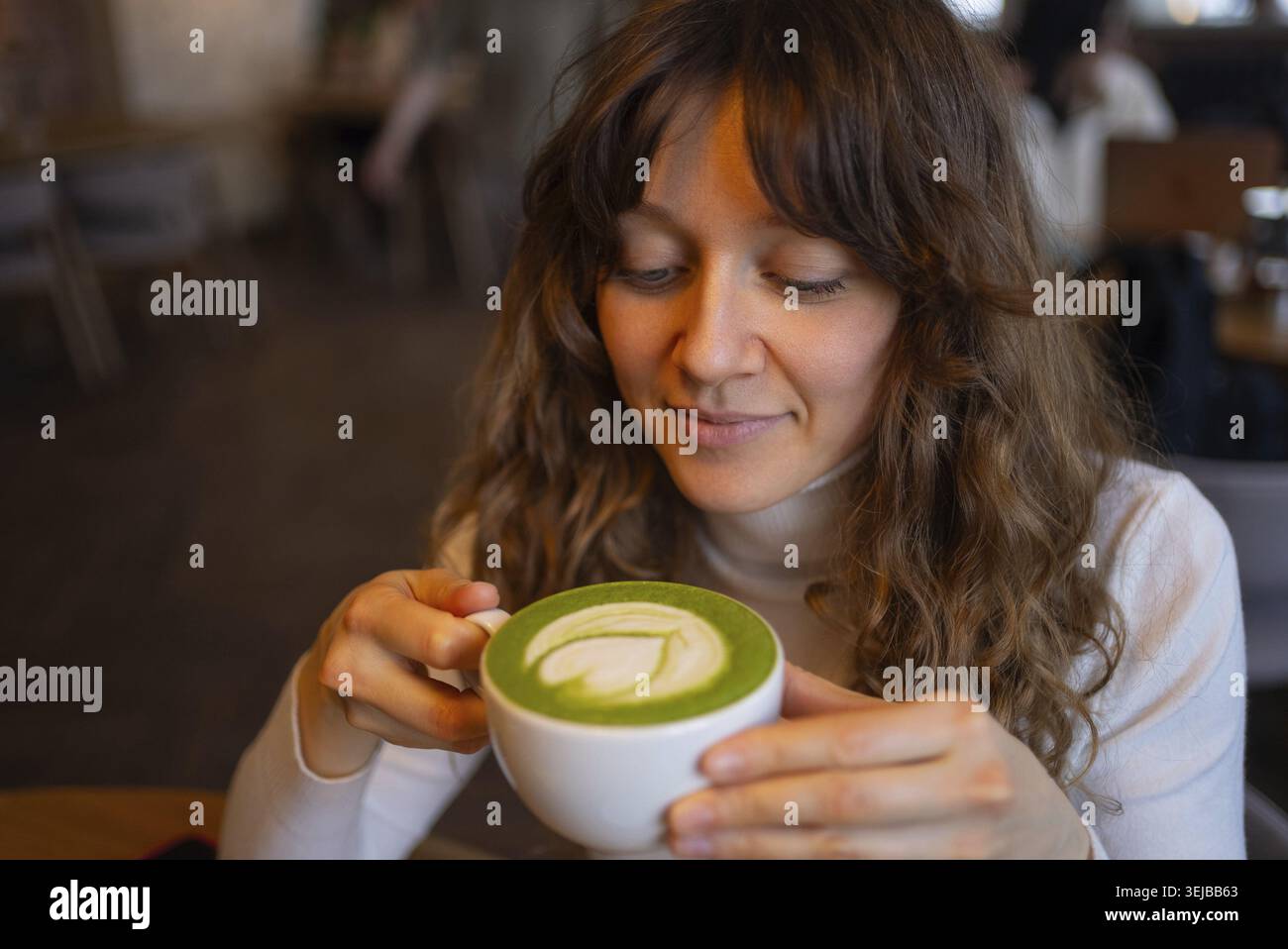 Junge Frau, die in einem gemütlichen Café einen warmen Matcha-Latte mit komplizierter Blattkunst genießt, genießt die friedliche Atmosphäre und die reichen, erdigen Aromen des Stockfoto