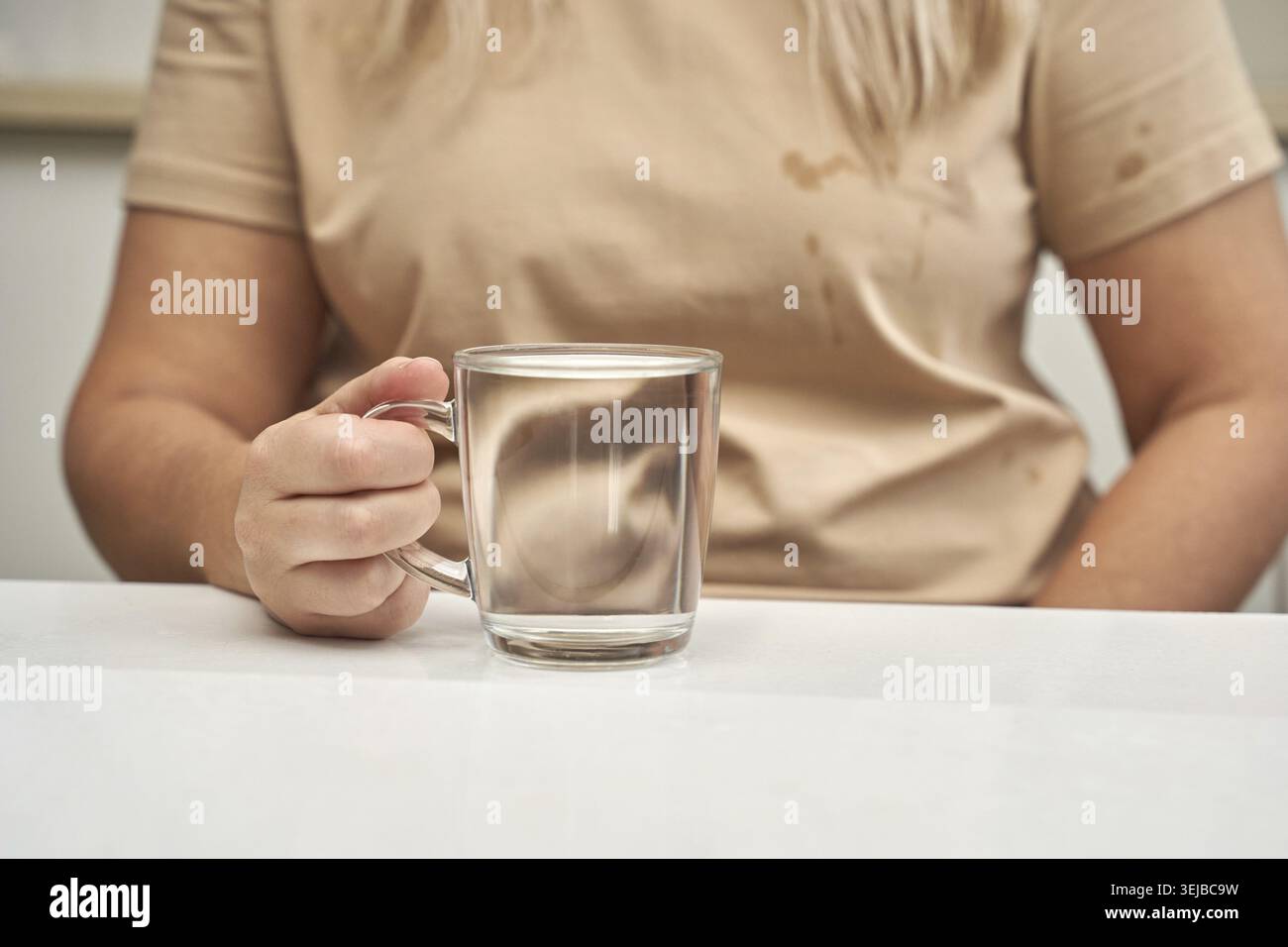 Weiblicher Erwachsener hält ein klares Glas Wasser an einem Tisch mit hellbeigem T-Shirt Stockfoto