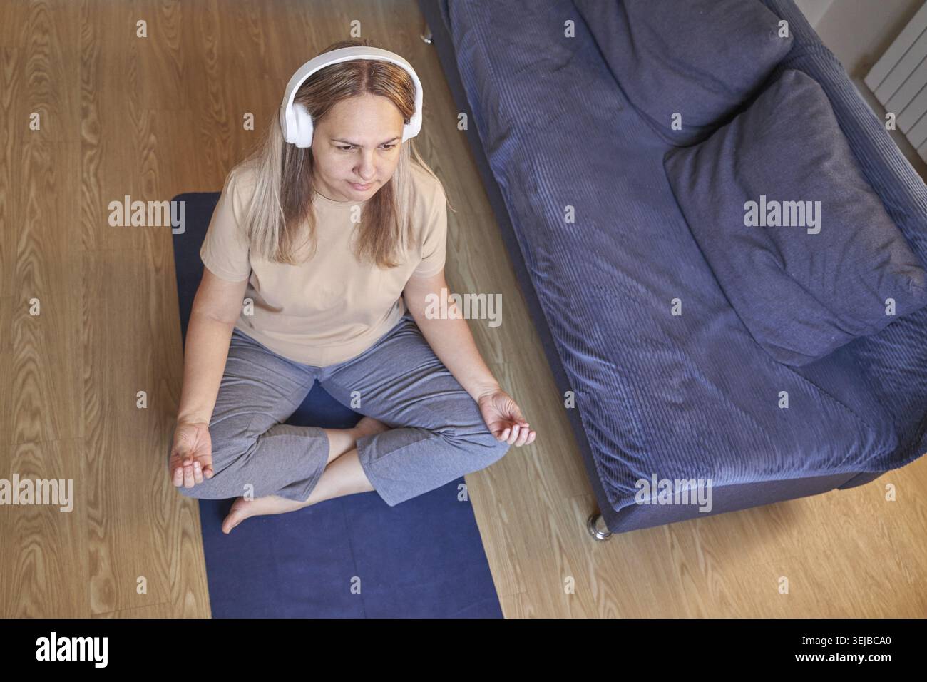 Reife weiße Frau übt zu Hause Meditation mit Kopfhörern, die auf dem Boden in der Nähe der Couch sitzen Stockfoto