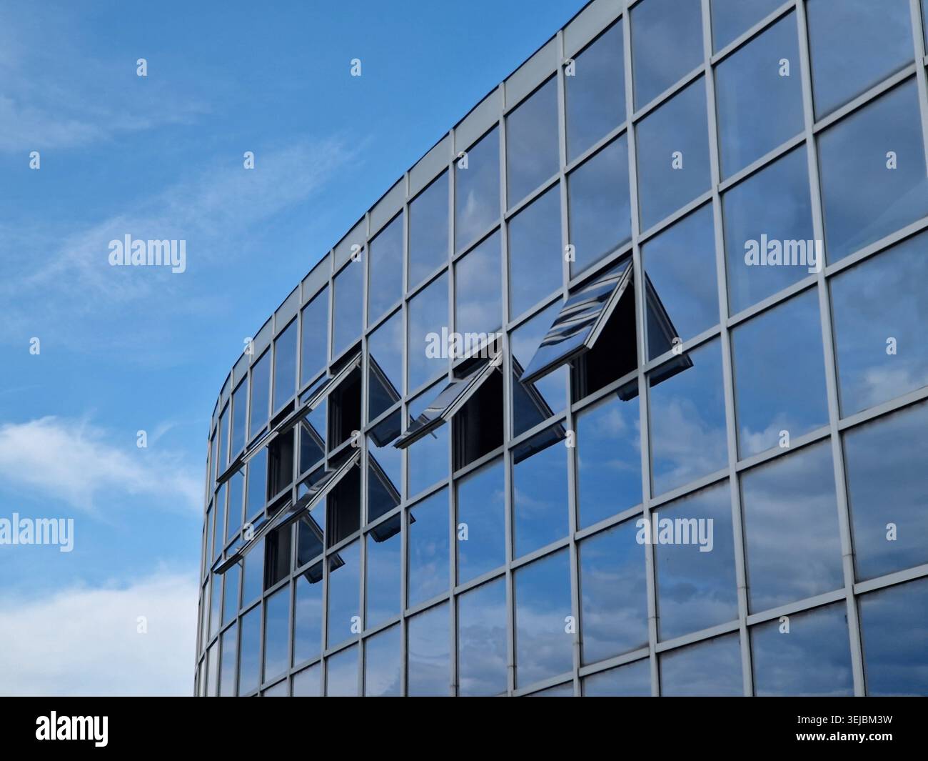 Geschwungenes Bürogebäude aus Glas reflektiert dramatisch bewölkten Himmel und Sonnenlicht. Moderne Unternehmensarchitektur mit blau getönten Fenstern, geometrischen Linien und Stockfoto