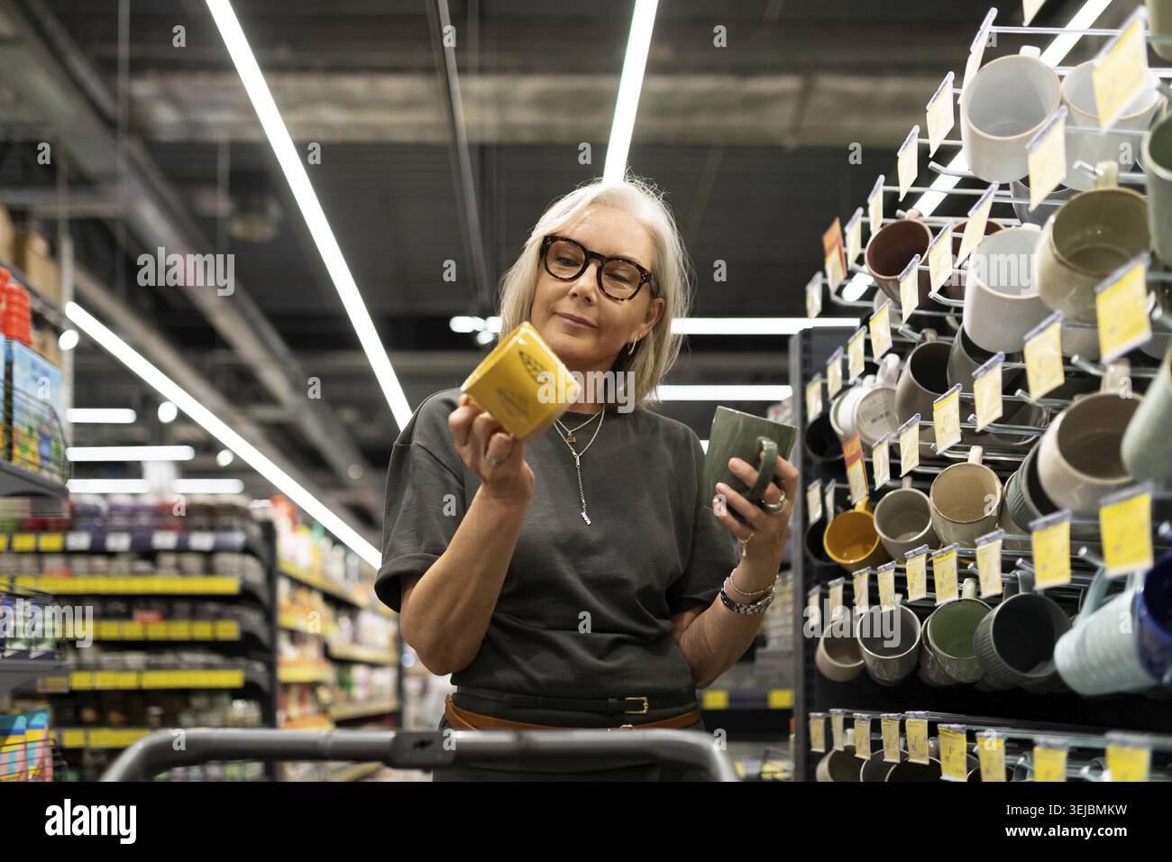 Eine stilvolle, reife Frau stöbert in einem modernen Geschäft für Haushaltswaren und wählt Pflanzgefäße und Küchenutensilien aus. Sie wählt nachdenklich Dinge für ihren Lebensunterhalt aus Stockfoto