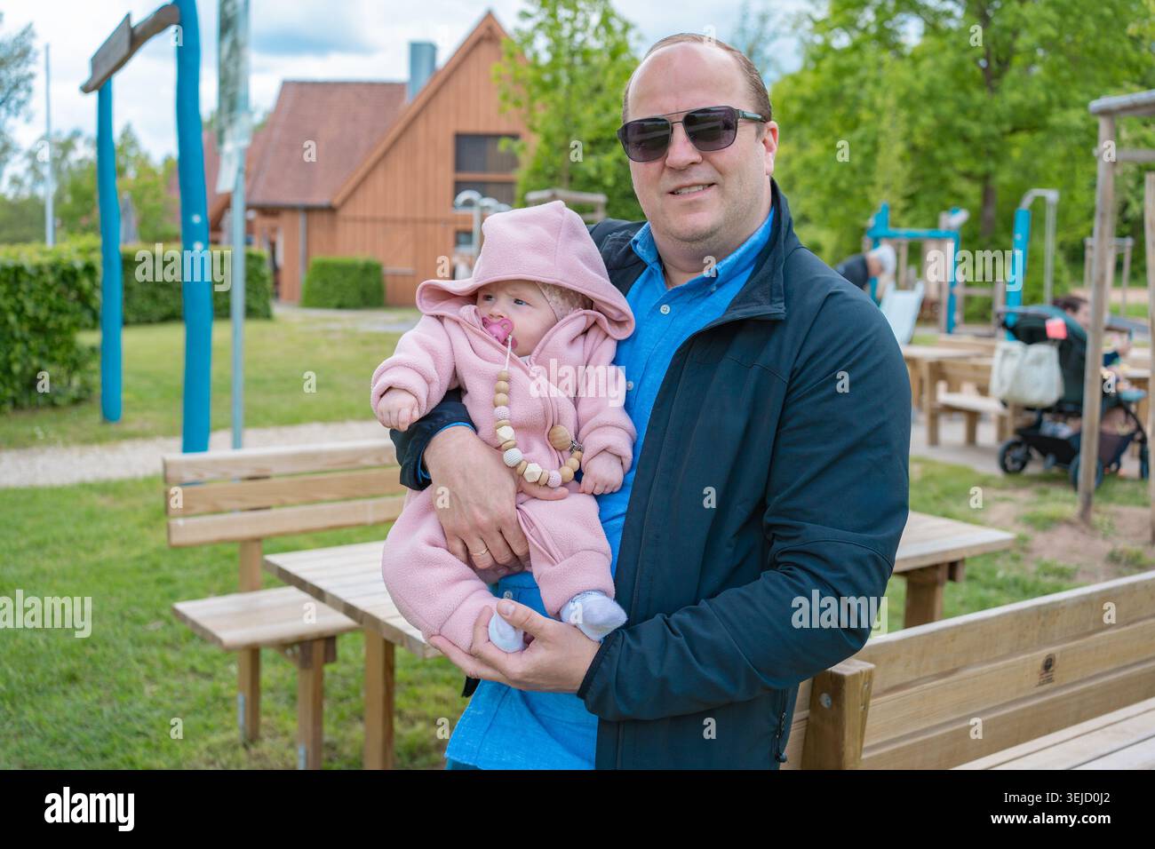 Ein glücklicher, liebevoller Vater mit Sonnenbrille hält sein kleines Kind in einem rosa warmen Jumpsuit auf einem öffentlichen Spielplatz im Freien. Das Konzept des Glücks Stockfoto