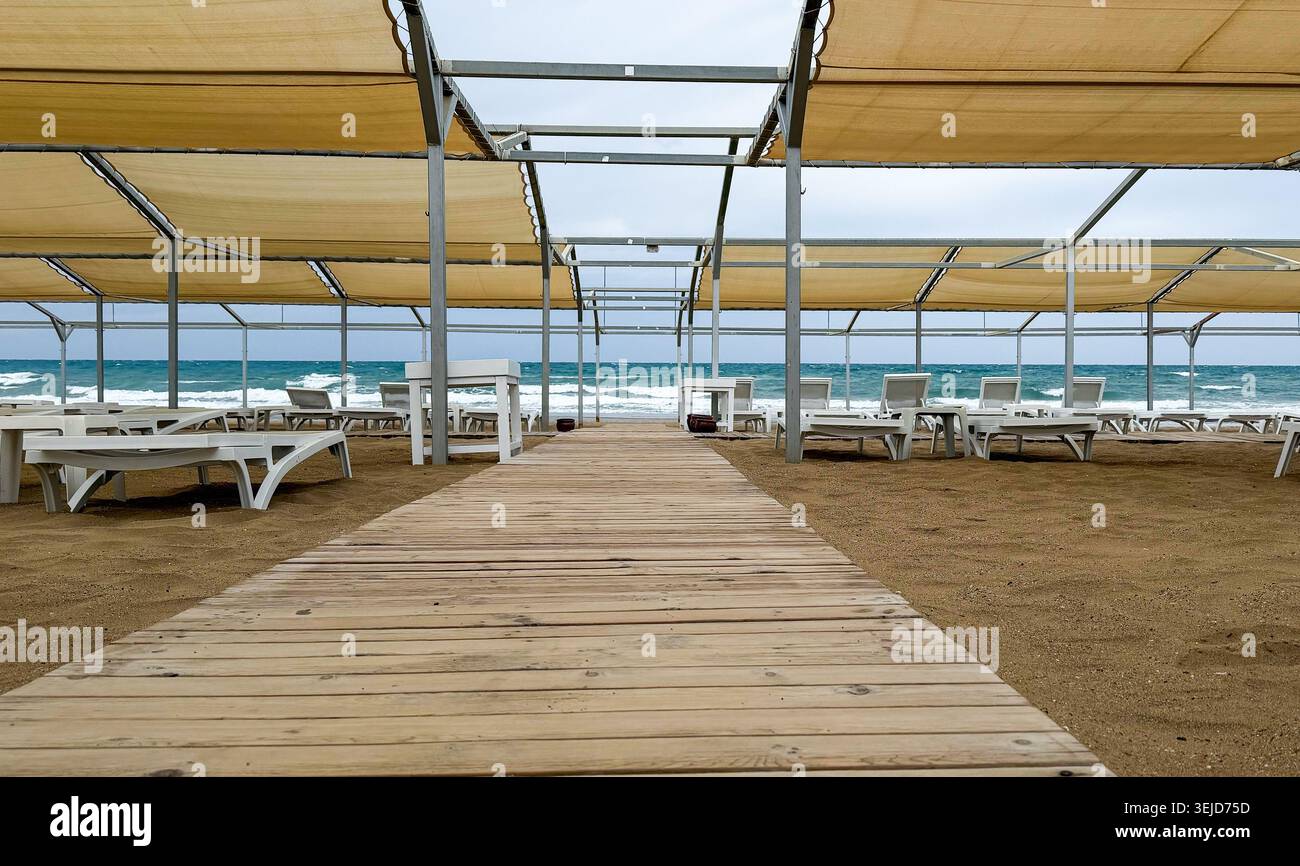 Hölzerner Pfad führt zum Lounge-Bereich am Strand mit weißen Sonnenliegen und Baldachinen, Ozeanwellen im Hintergrund unter bewölktem Himmel sichtbar Stockfoto
