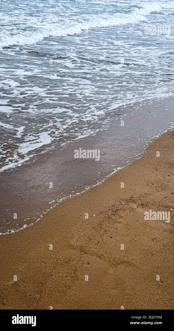 Die Wellen schlagen sanft gegen einen Sandstrand, mit einer klaren Küstenlinie und Schaumbildung entlang des Wasserrandes, die eine ruhige Küstenszene schaffen Stockfoto
